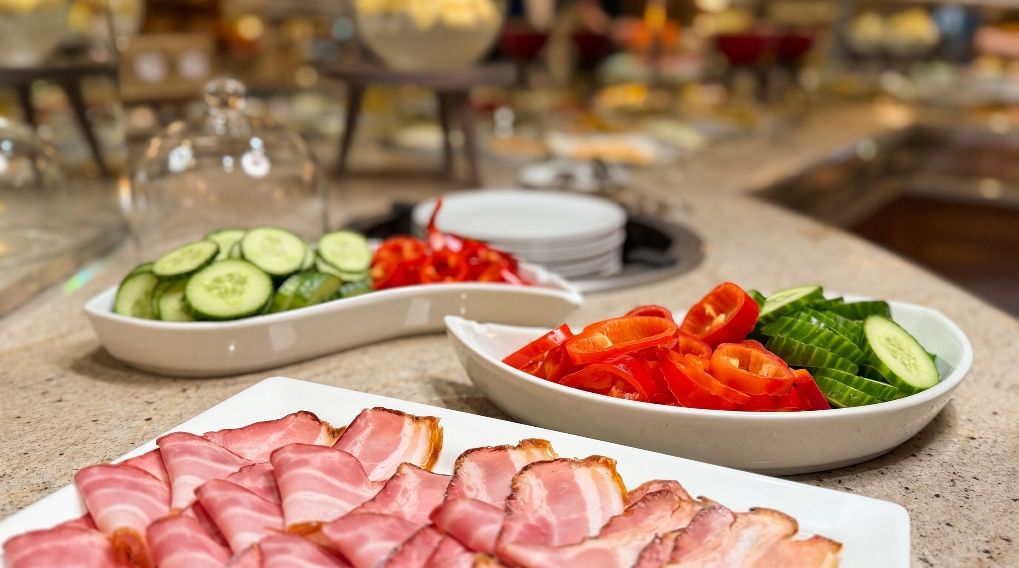 A buffet table with sliced meats, vegetables, and a glass container.