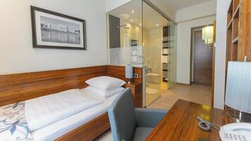 A hotel room with a wooden bed, a desk with a telephone, a chair, a glass partition, and a framed picture on the wall.