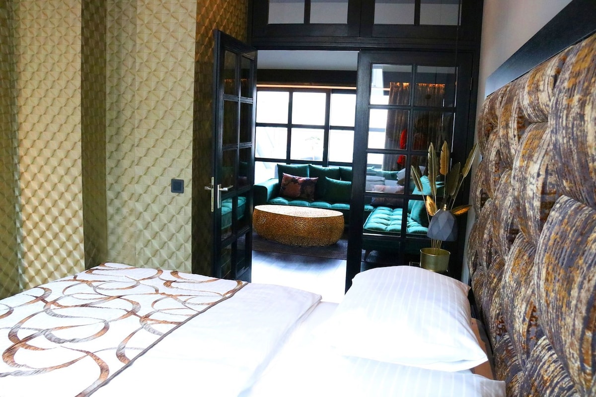 A bedroom with a bed, a green sofa, a wicker ottoman, and a view through an open door.