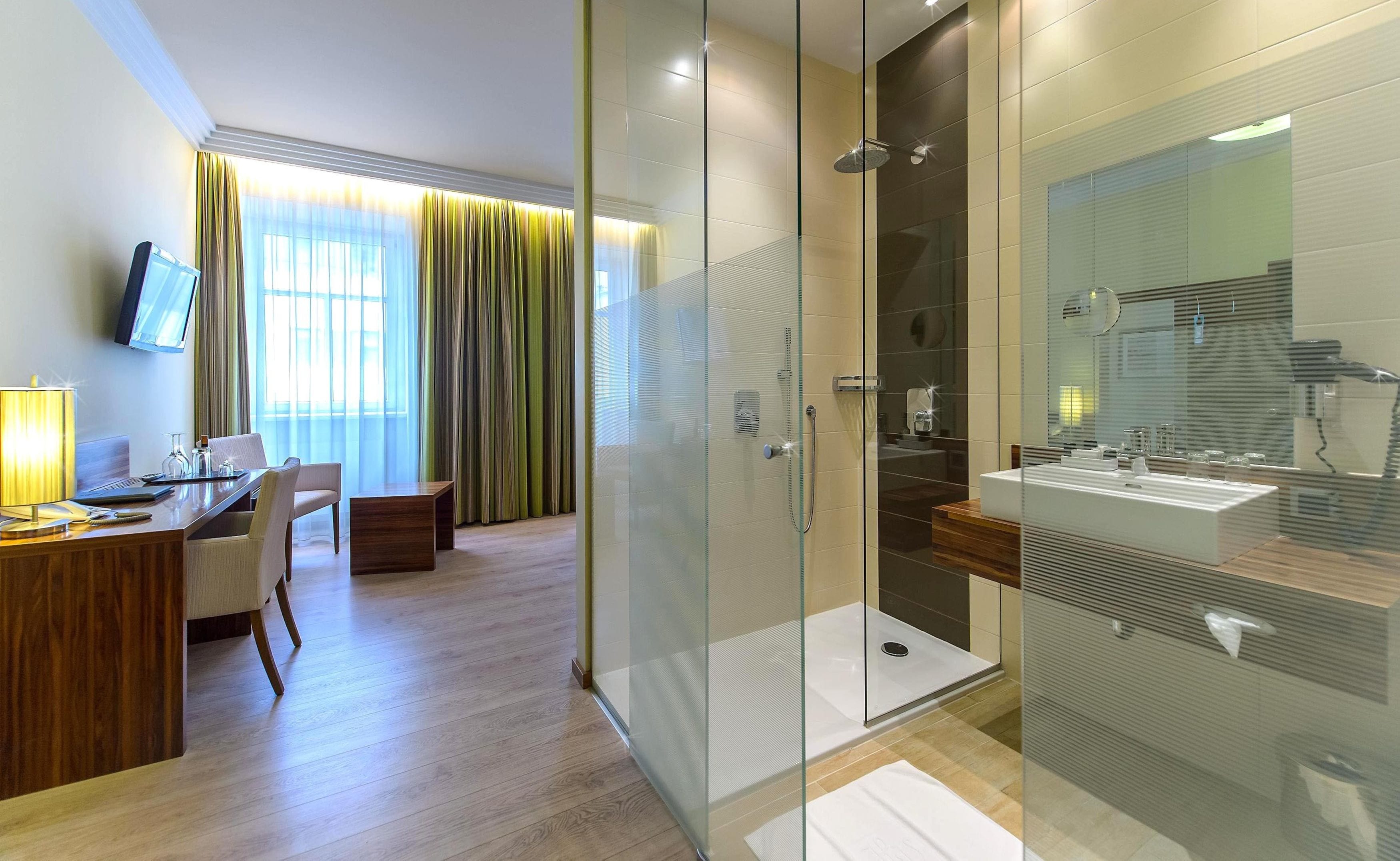 Ein modernes Hotelzimmer mit einer gläsernen Dusche, einem Holztisch mit Stuhl und einem großen Fenster mit Vorhängen.