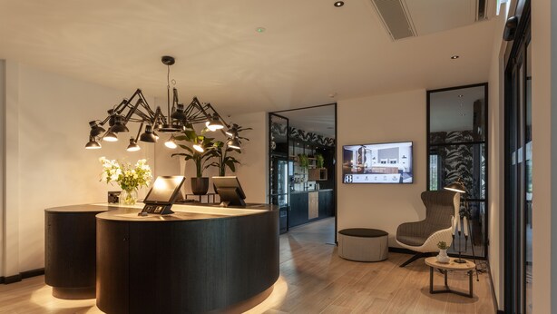 A modern reception area with a curved desk, multiple pendant lights, a flat-screen TV, and a comfortable seating area with a chair and a small table. - Gr8 Hotel Sevenum (Sevenum)