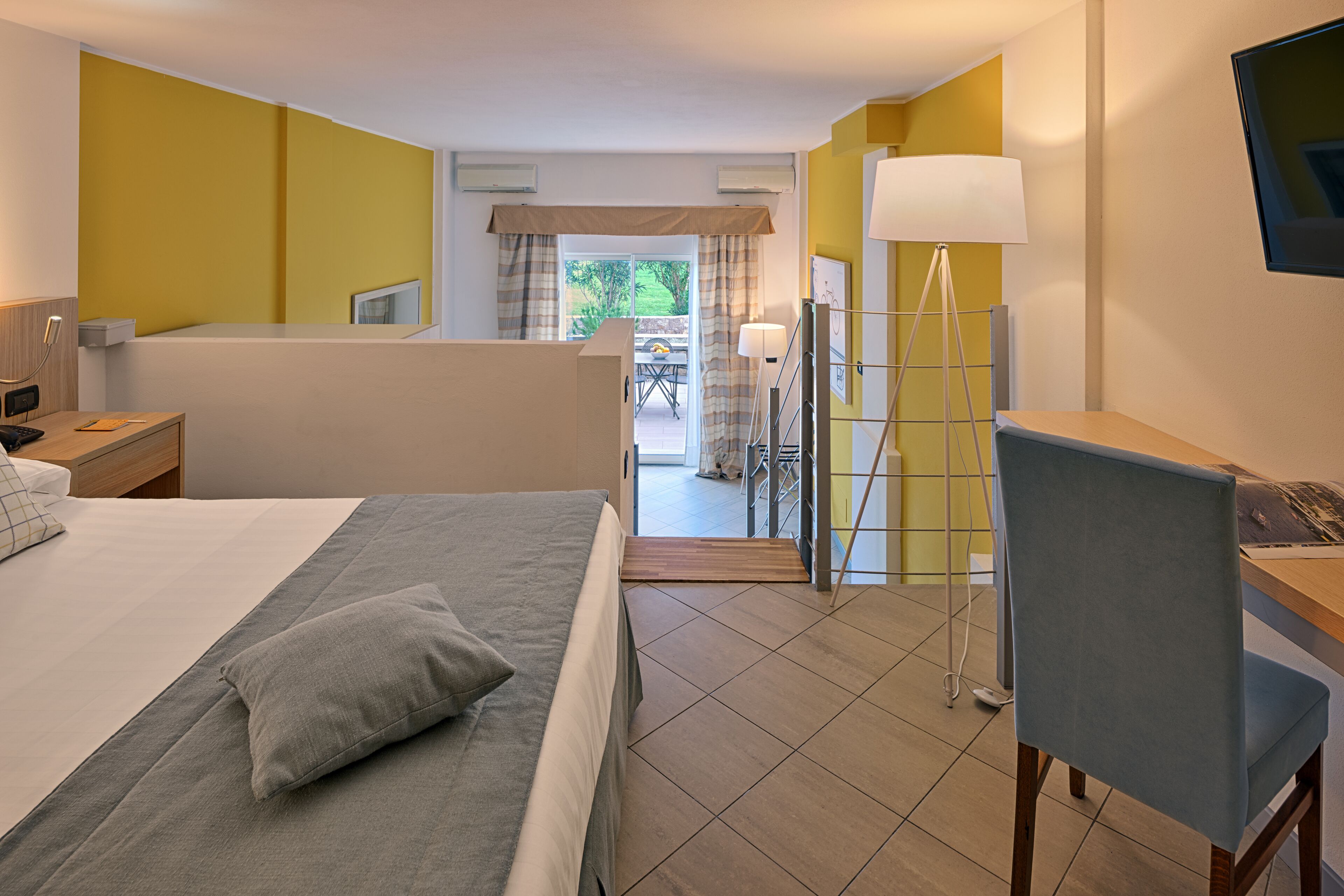 A hotel room with a bed, a desk, a chair, a lamp, and a view of the outside.