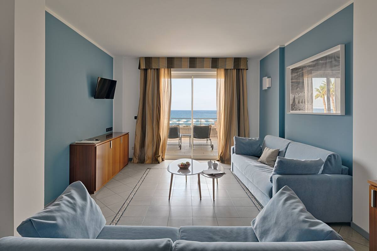 A modern living room with a sofa, a coffee table, and a TV on a wooden cabinet. A balcony with a view of the sea is visible through large windows.