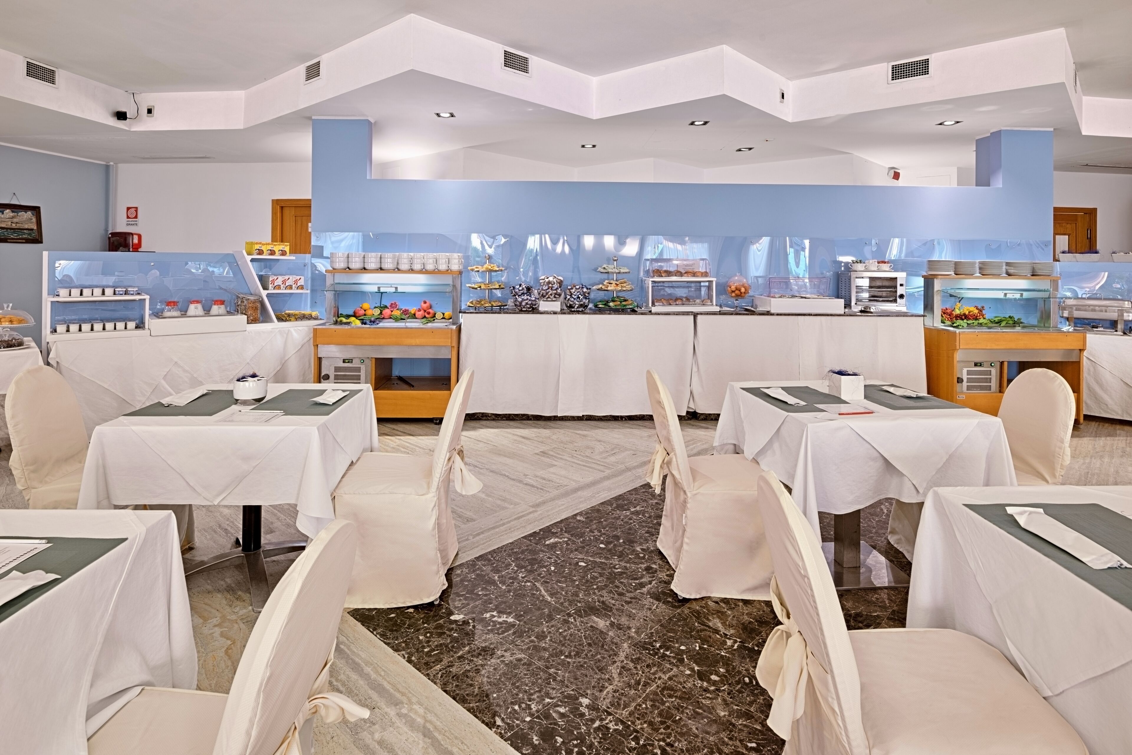 Un comedor de hotel con varias mesas y sillas, un bufé con diversos alimentos y un piso de mármol.