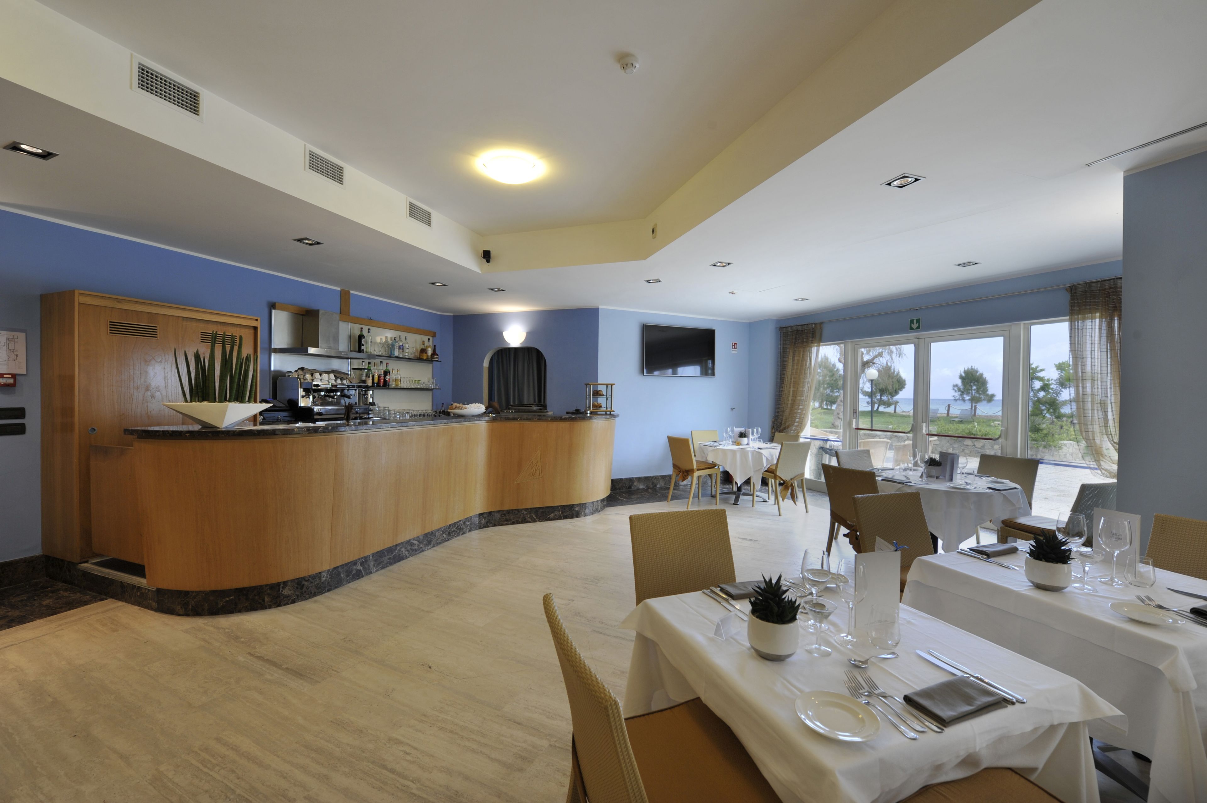 Zona de comedor de hotel con una barra de madera curvada, mesas y sillas blancas.