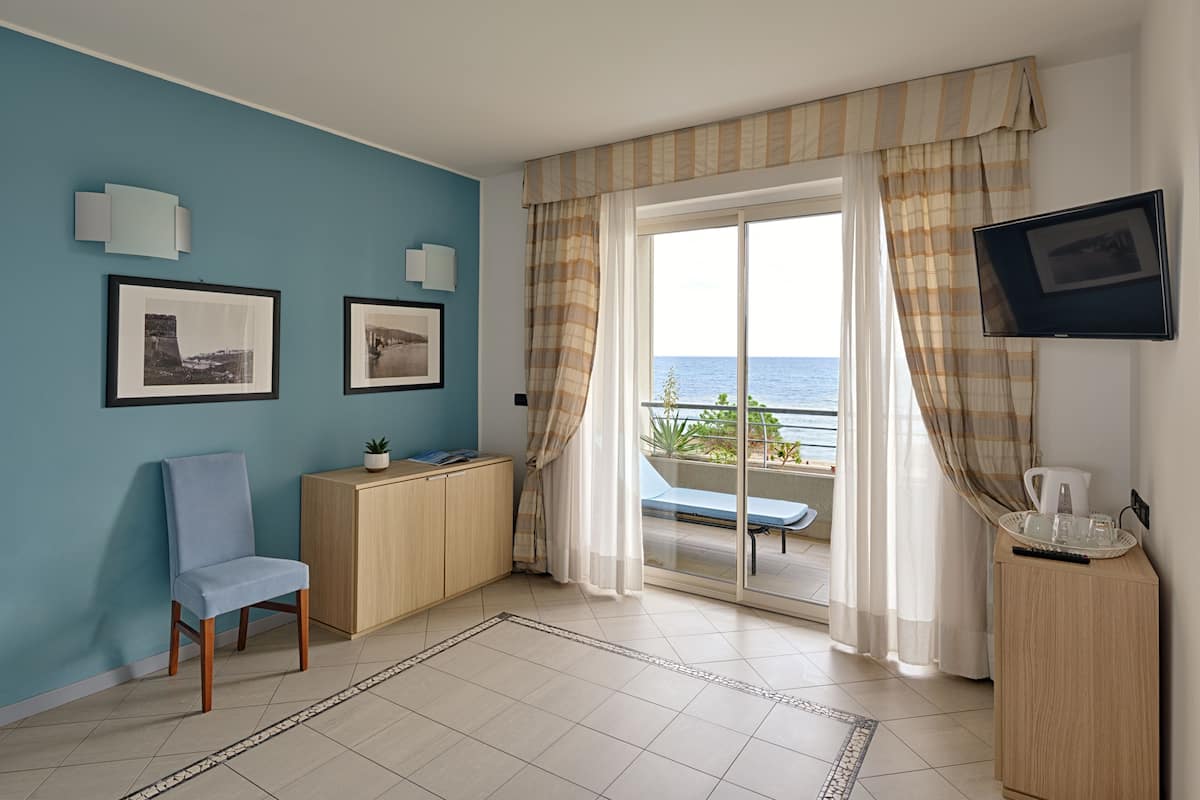 A room with a blue wall, a chair, a small cabinet, a television, and a view of the sea through a large window with curtains.