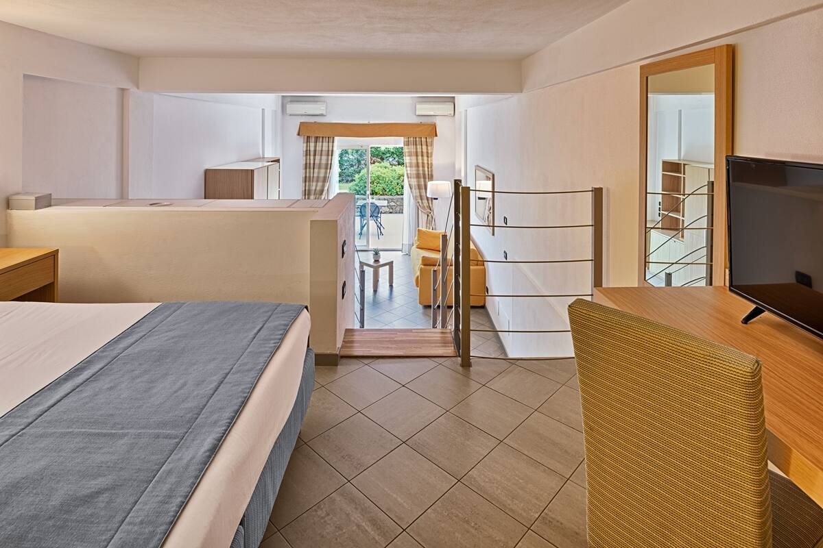 A hotel room with a bed, a desk, a chair, a TV, and a view of the outdoor area.
