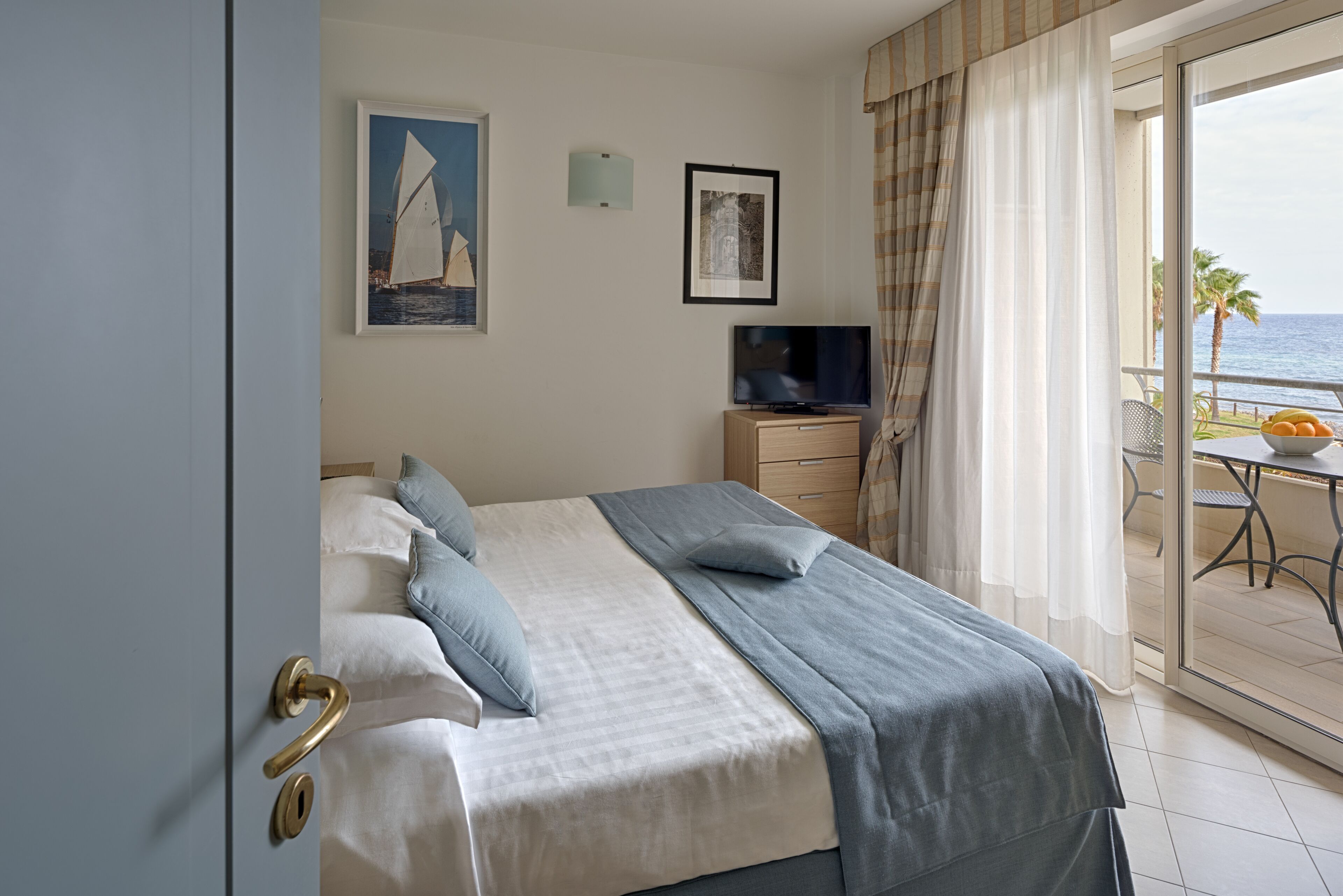 A hotel room with a bed, a TV, and a balcony with a view of the sea.