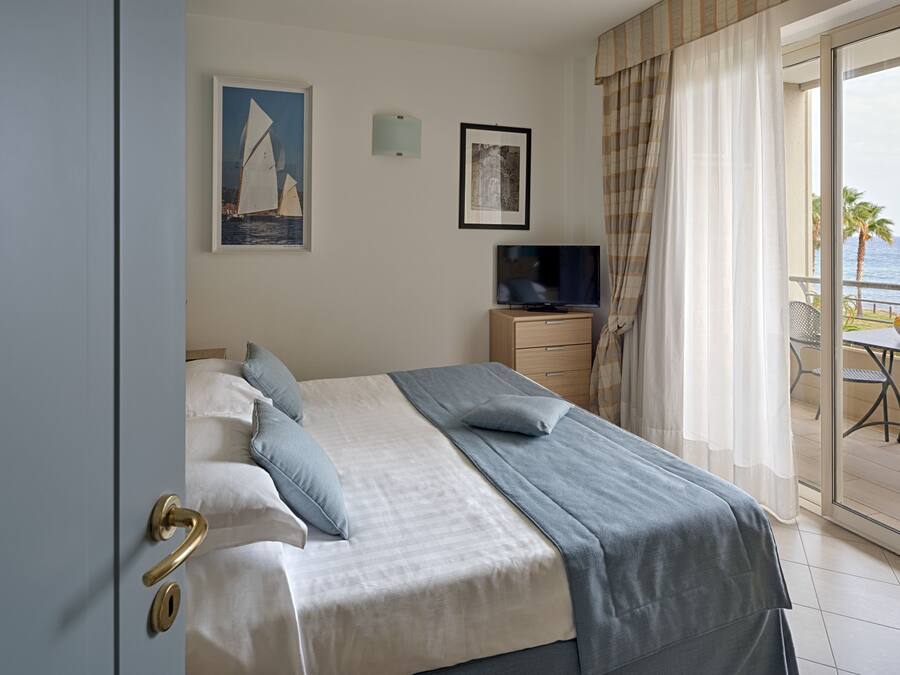 A hotel room with a bed, a TV, and a balcony with a view of the sea.
