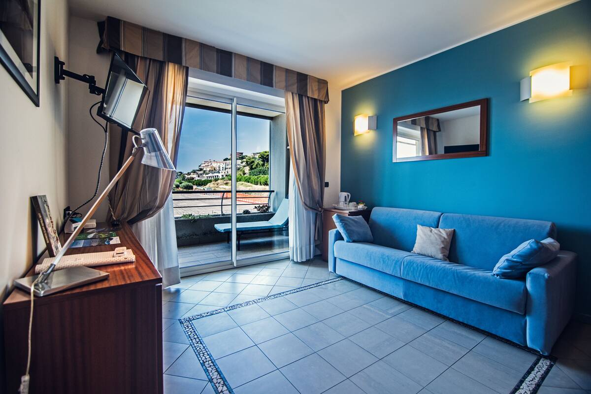A modern hotel room with a blue sofa, a desk with a laptop, a large window with curtains, and a view of buildings and greenery.