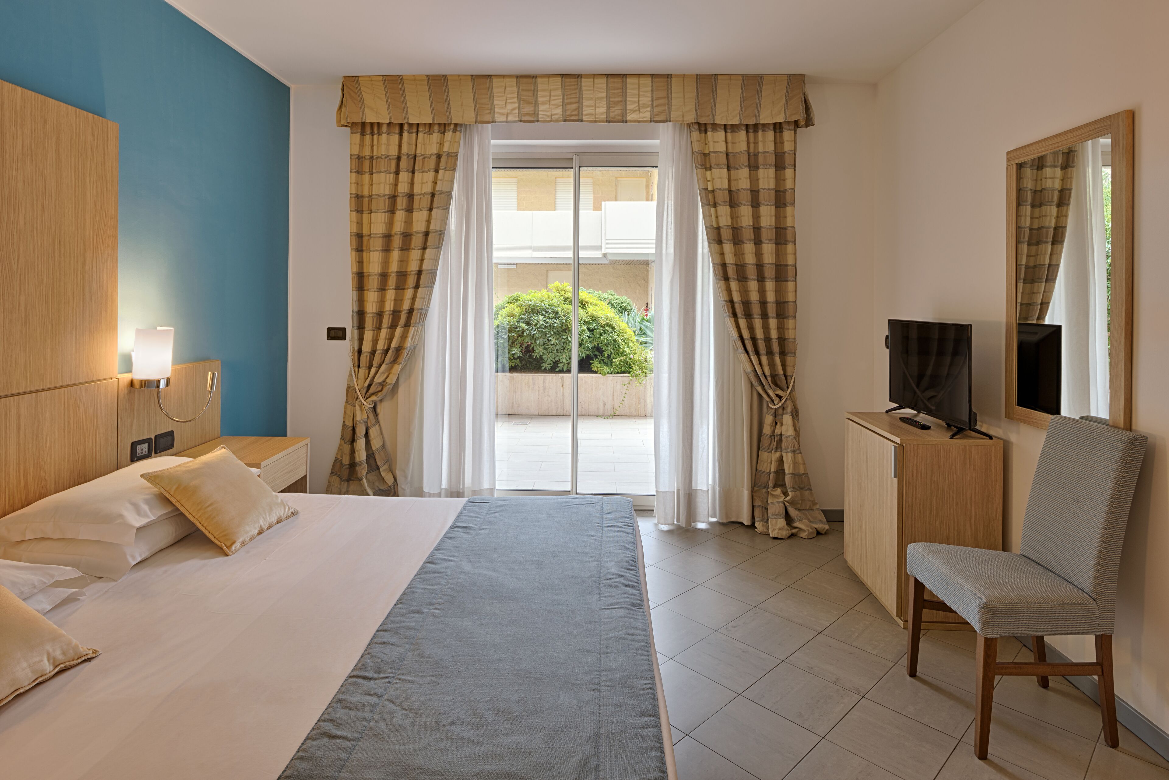 A hotel room with a bed, a desk with a TV, a chair, and a window with curtains.