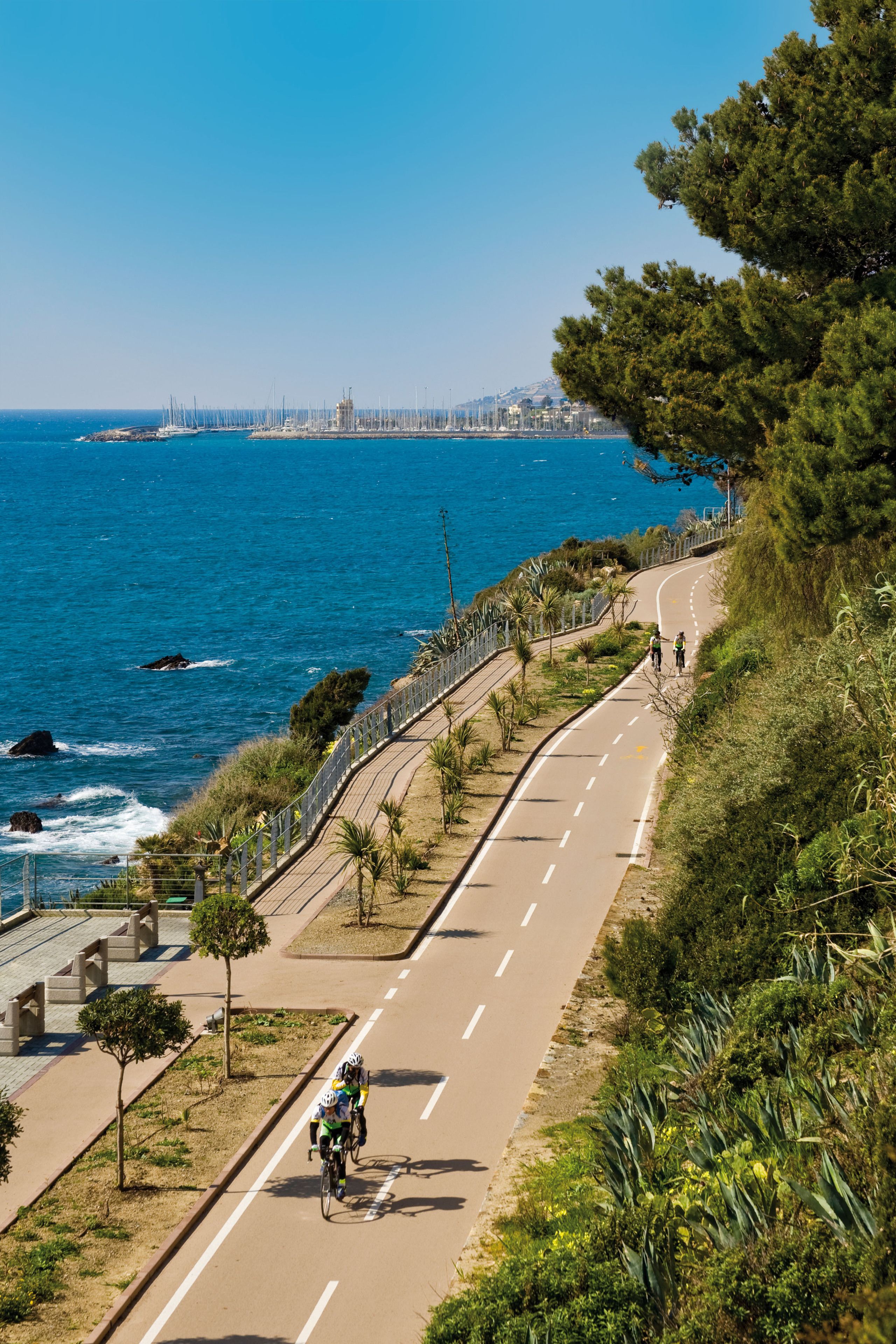 A coastal road with a cyclist, a clear blue sea, and a cityscape in the background.