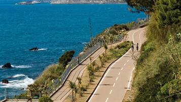A coastal road with a cyclist, a clear blue sea, and a cityscape in the background.
