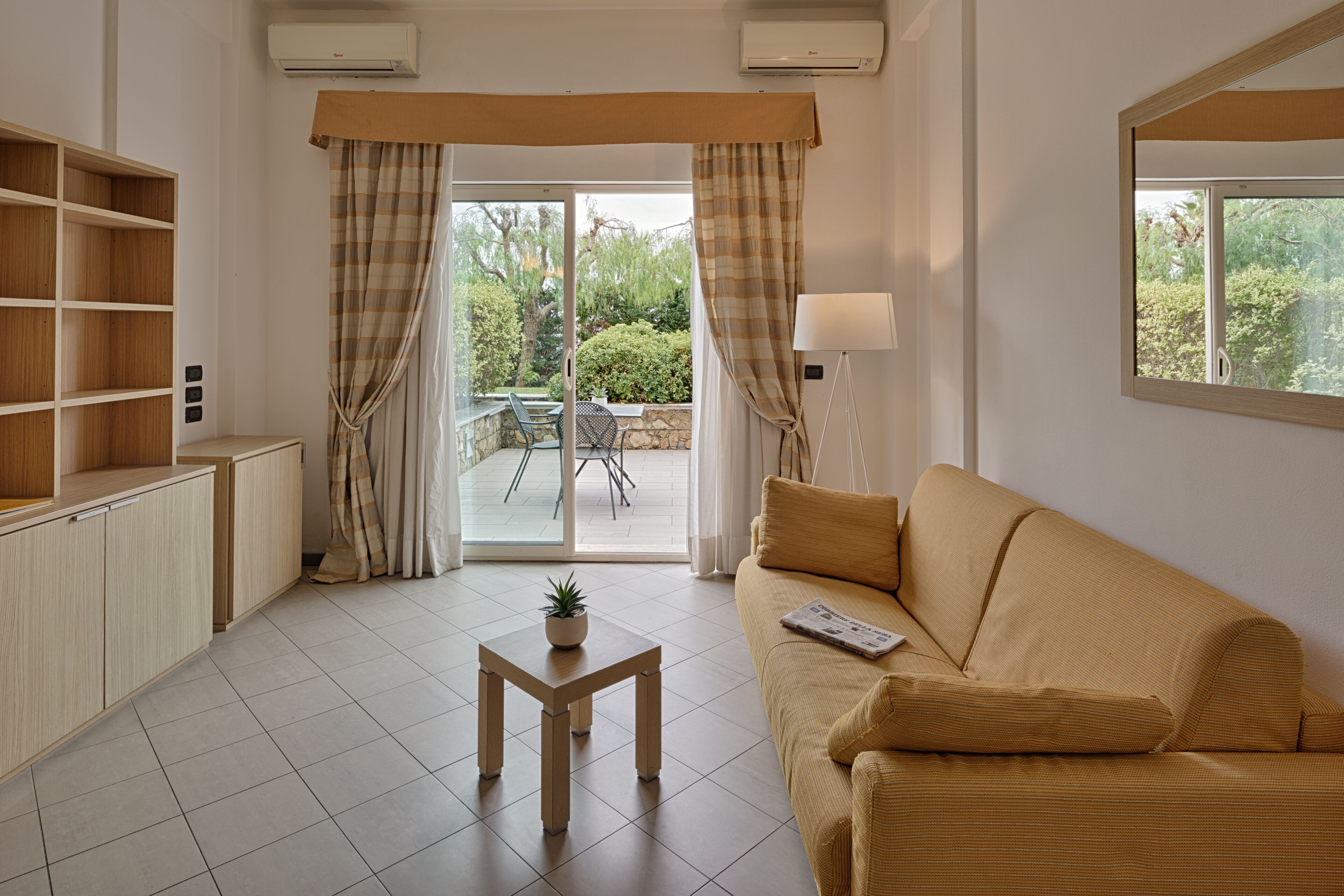 Una sala de estar con un sofá, una mesita con una planta, un mueble de madera y un ventanal corredizo que da a un área exterior con una mesa y sillas.