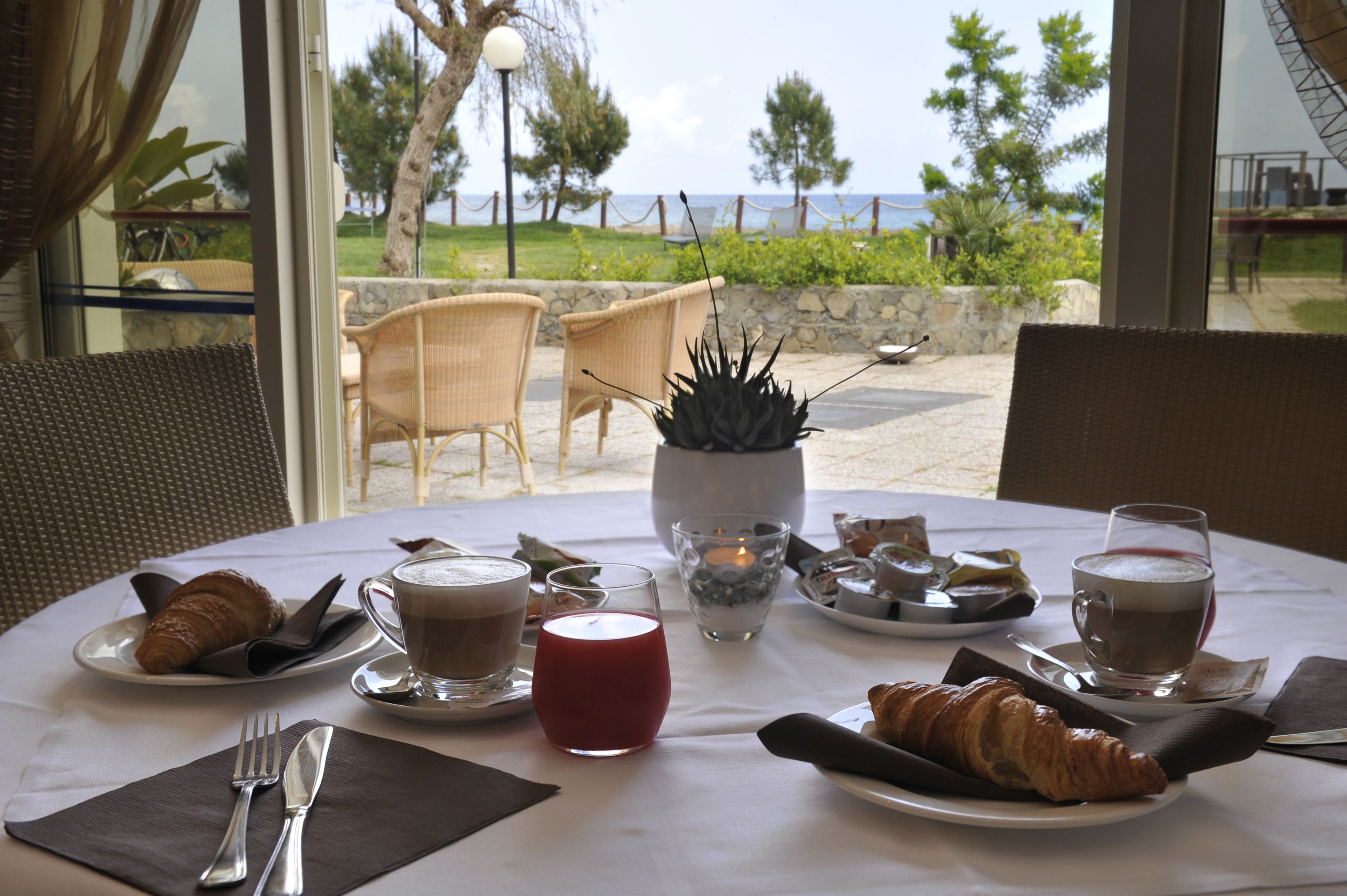 Une table dressée pour le petit-déjeuner, avec des croissants, du café et une boisson rouge, offrant une vue magnifique sur la mer et la verdure environnante.