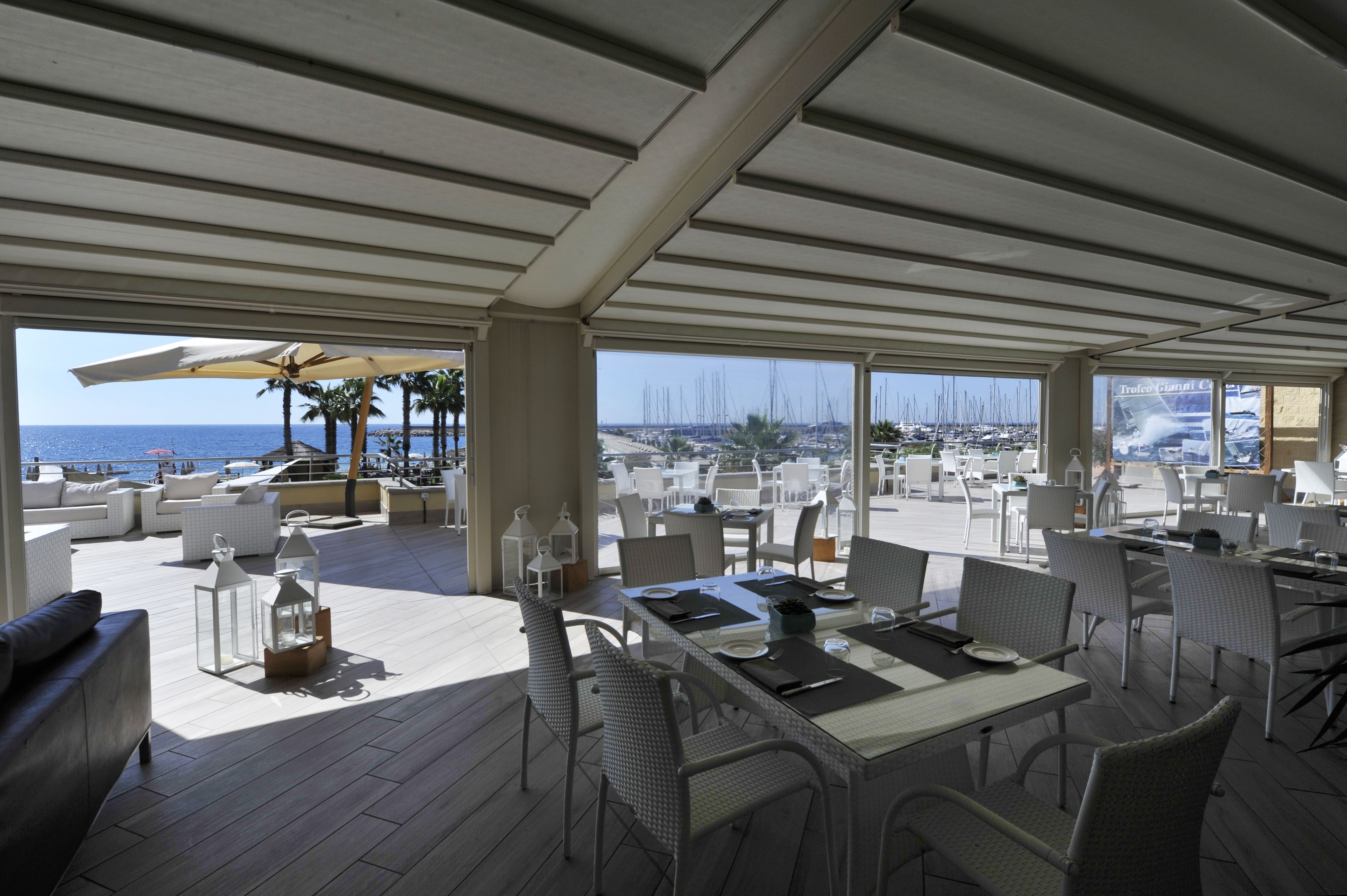An outdoor dining area with tables and chairs, a view of the sea, and a covered section with umbrellas.