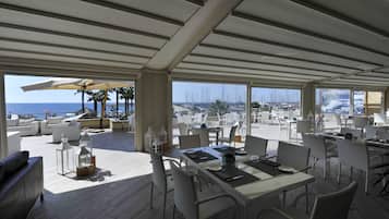 Un área de comedor al aire libre con mesas y sillas, vista al mar y una sección cubierta con sombrillas.