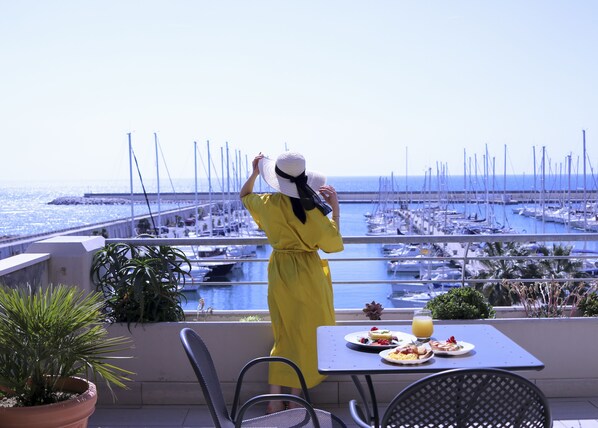 Una mujer con vestido amarillo y sombrero blanco observa un puerto deportivo con barcos, una mesa con desayuno y plantas en macetas.