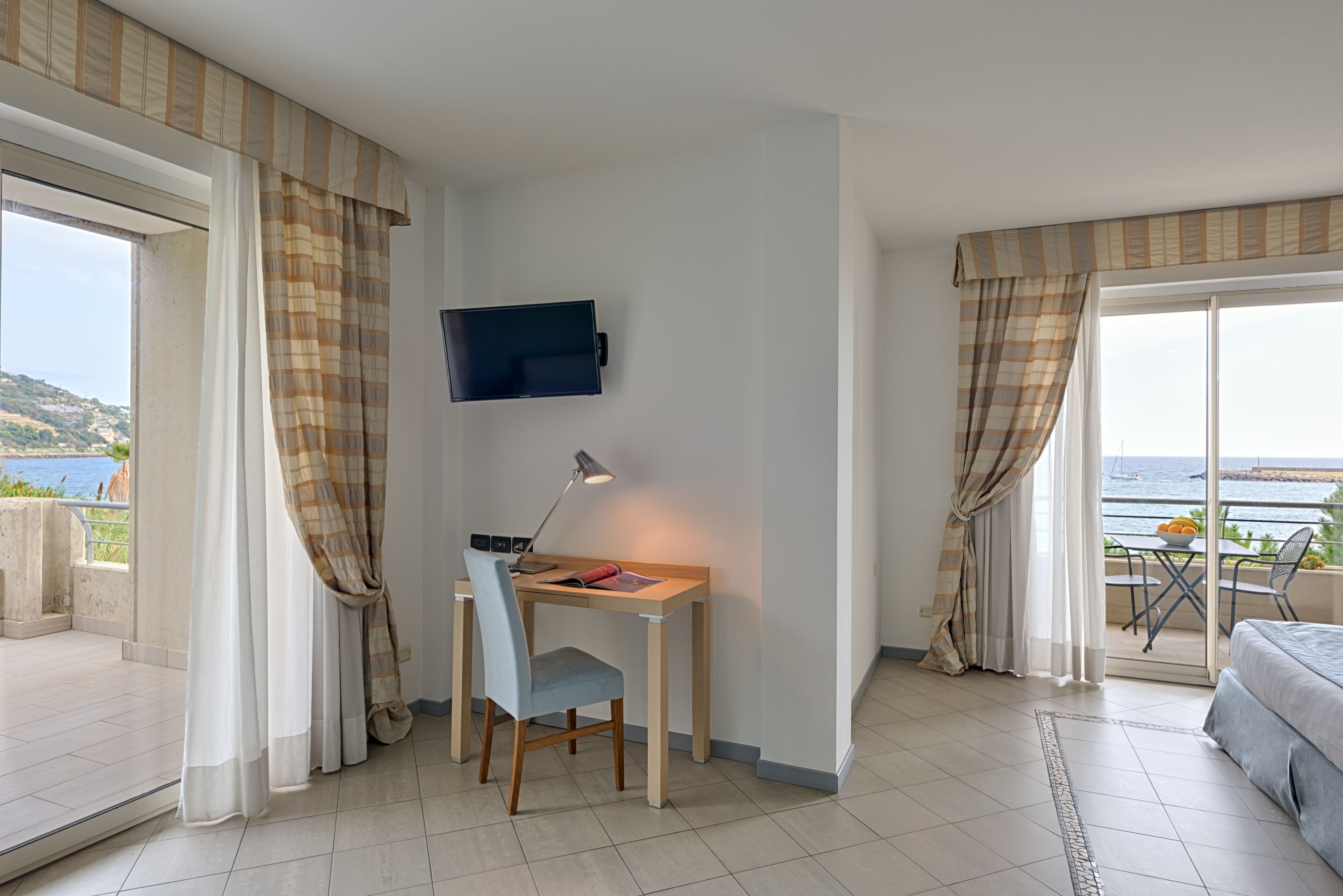 A hotel room with a desk, chair, and a TV mounted on the wall. There is a bed and a balcony with a view of the sea.