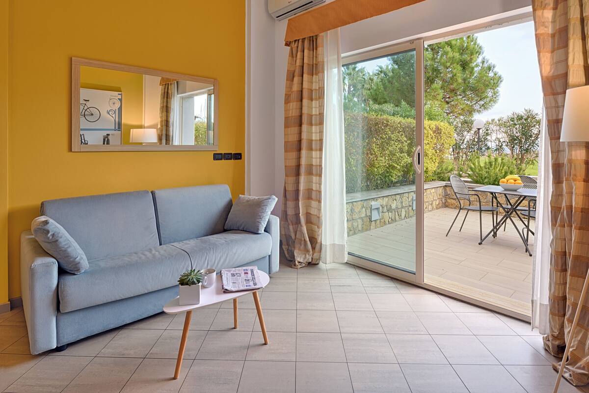 A modern living room with a sofa, a small table with a plant and newspaper, a large sliding glass door leading to an outdoor patio with a table and chairs.