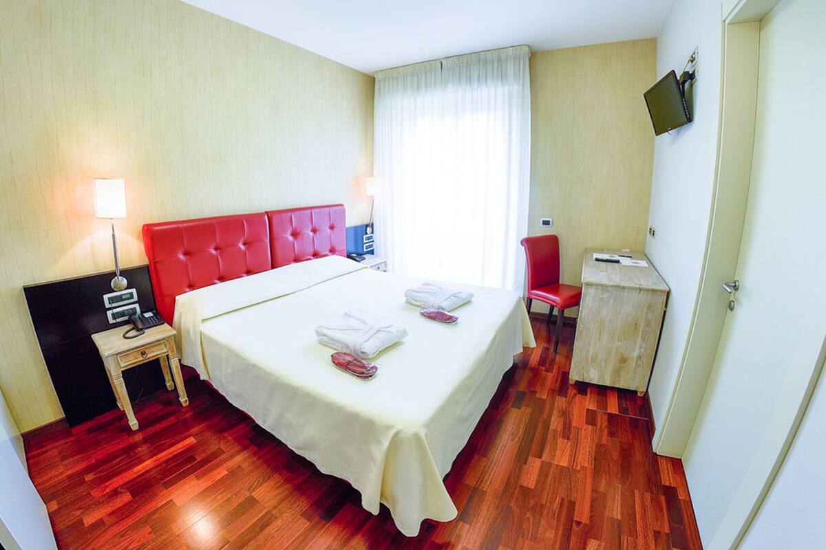 A hotel room with a large bed, a desk, a chair, a lamp, and a television on the wall.