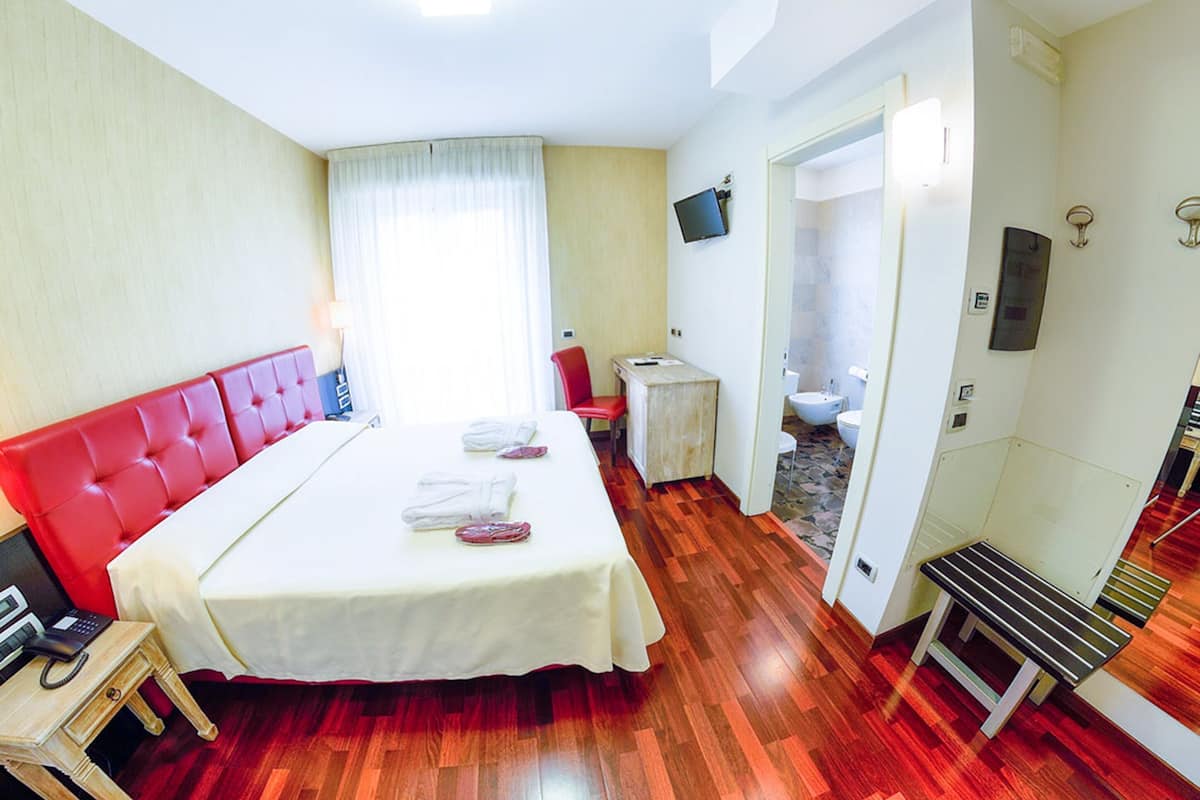 A hotel room with a red upholstered headboard, a bed with white linens, a small bedside table, a television mounted on the wall, and a wooden bench.