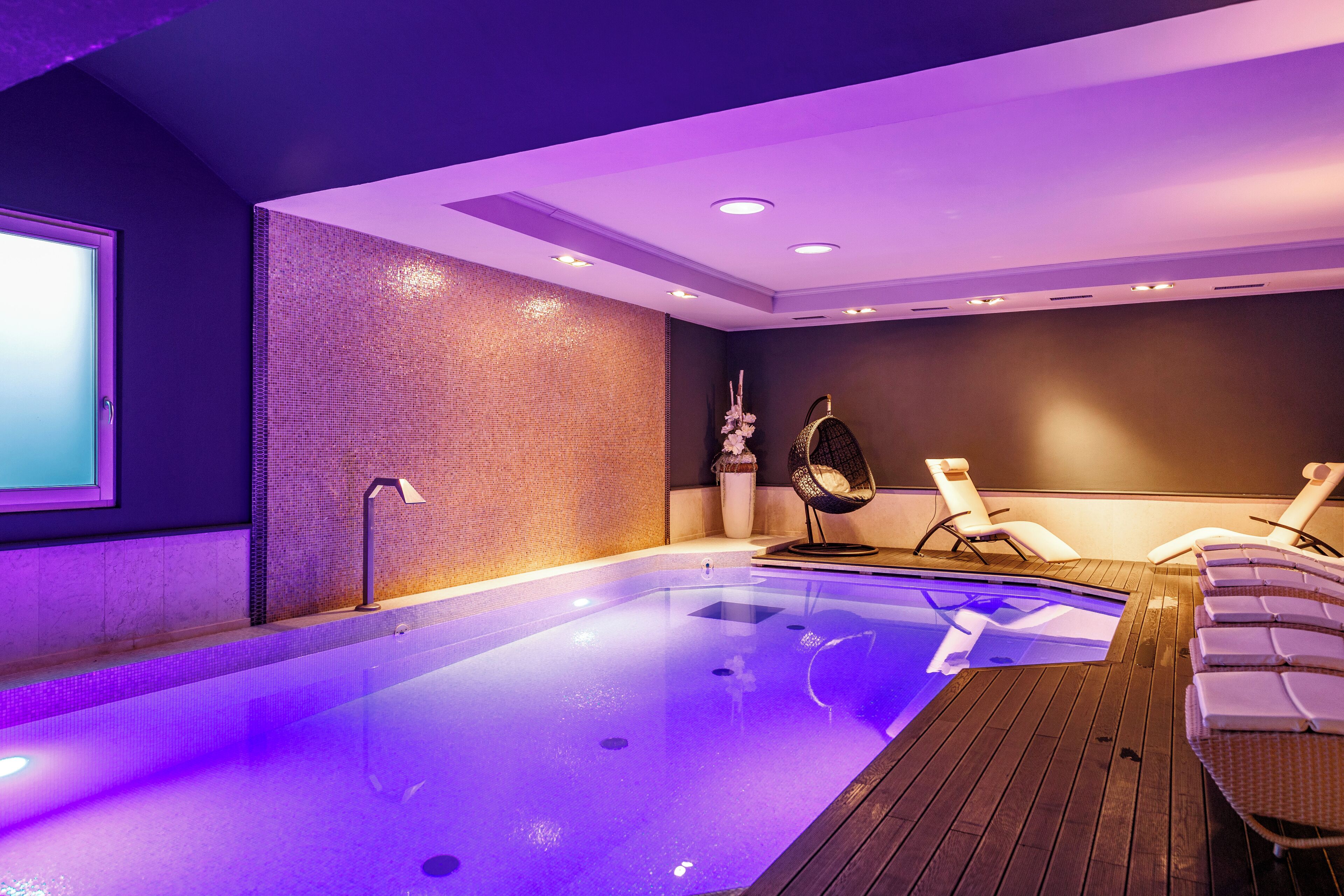 Un área de piscina interior moderna con un piso de madera, sillones de descanso y una pantalla grande en la pared.