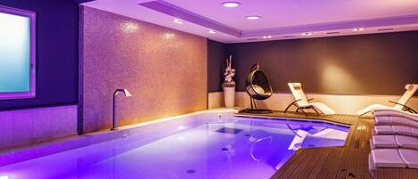 Un área de piscina interior moderna con un piso de madera, sillones de descanso y una pantalla grande en la pared.