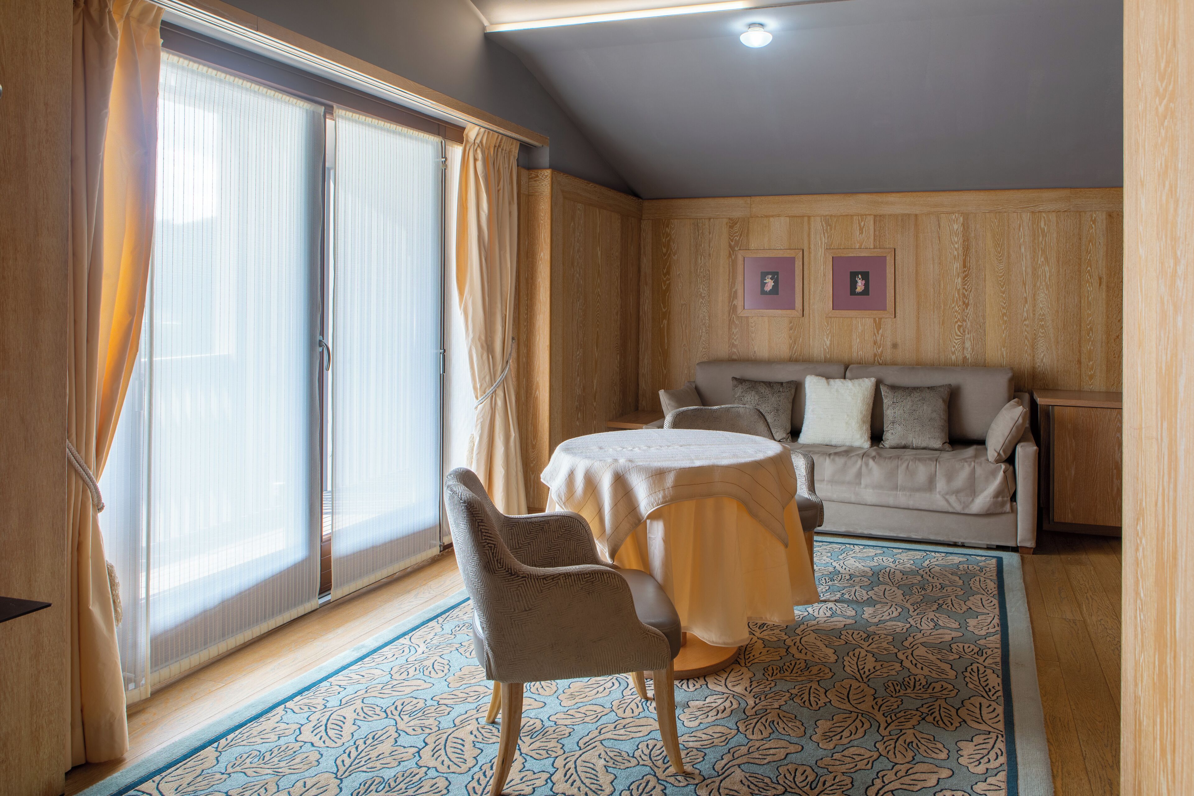 Une pièce dotée d’une porte-fenêtre coulissante, d’une table ronde avec une chaise, d’un canapé et de deux tableaux encadrés accrochés au mur.