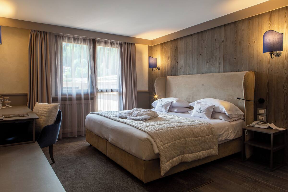 Ein Hotelzimmer mit einem großen Bett, einem Schreibtisch und einem Stuhl. Vor den Fenstern hängen Vorhänge und an der Wand ist eine Lampe befestigt.