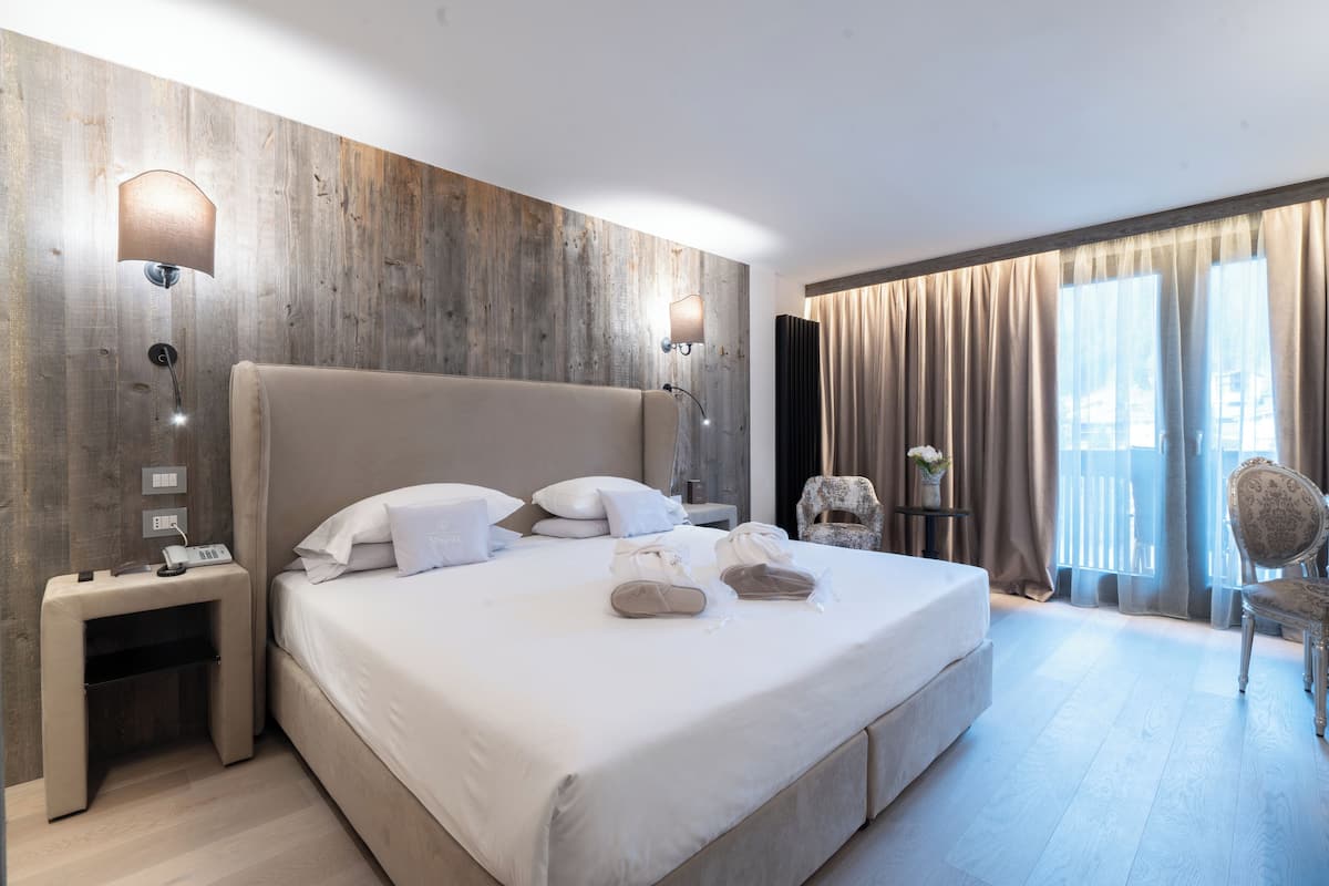 Ein Hotelzimmer mit einem großen Bett, zwei Nachttischlampen, einem Nachttisch, einem Sessel und Blick ins Freie durch ein Fenster mit Vorhängen.