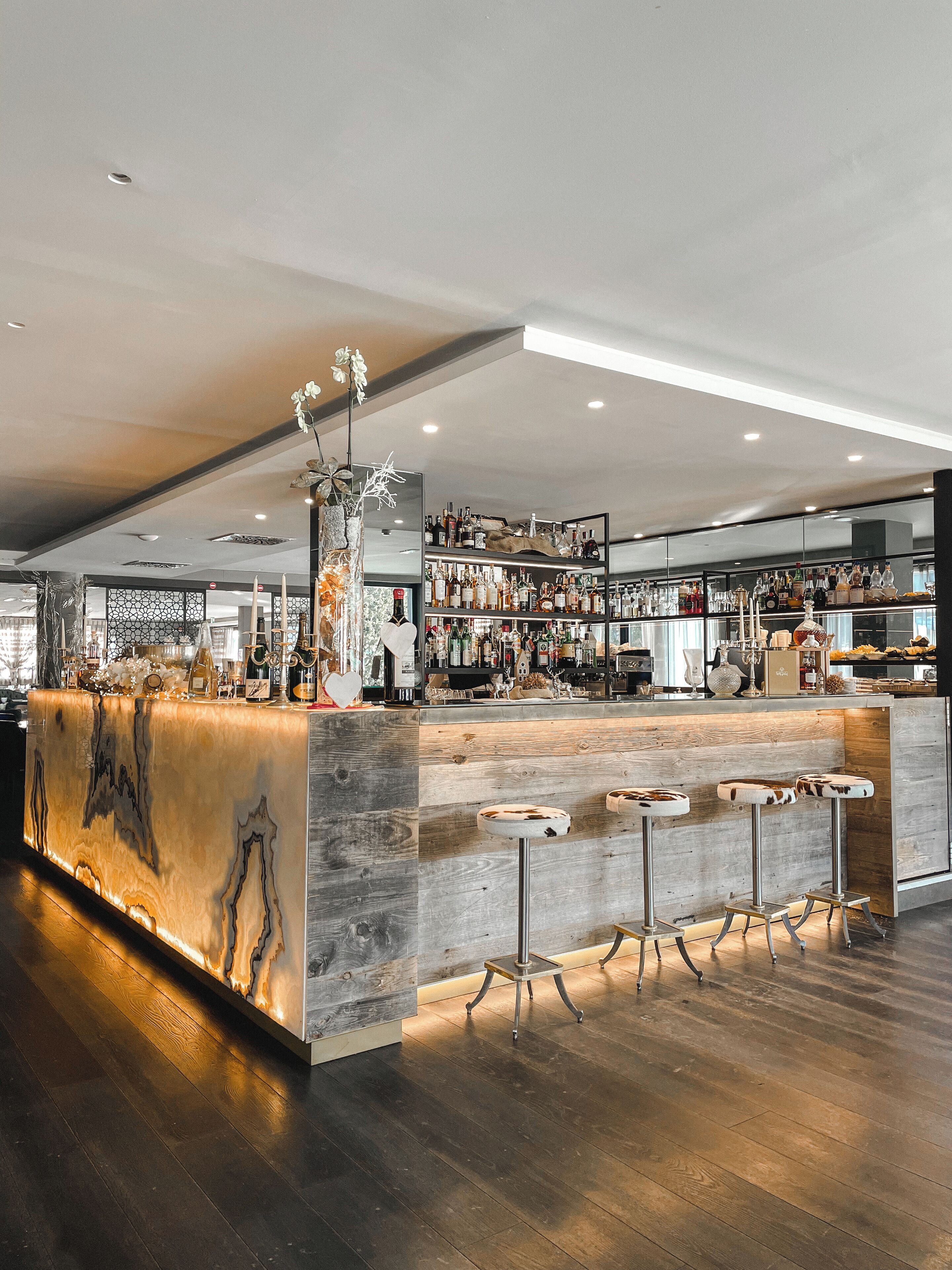 A modern bar area with a wooden bar counter, multiple stools, and a variety of bottles and glasses on shelves.