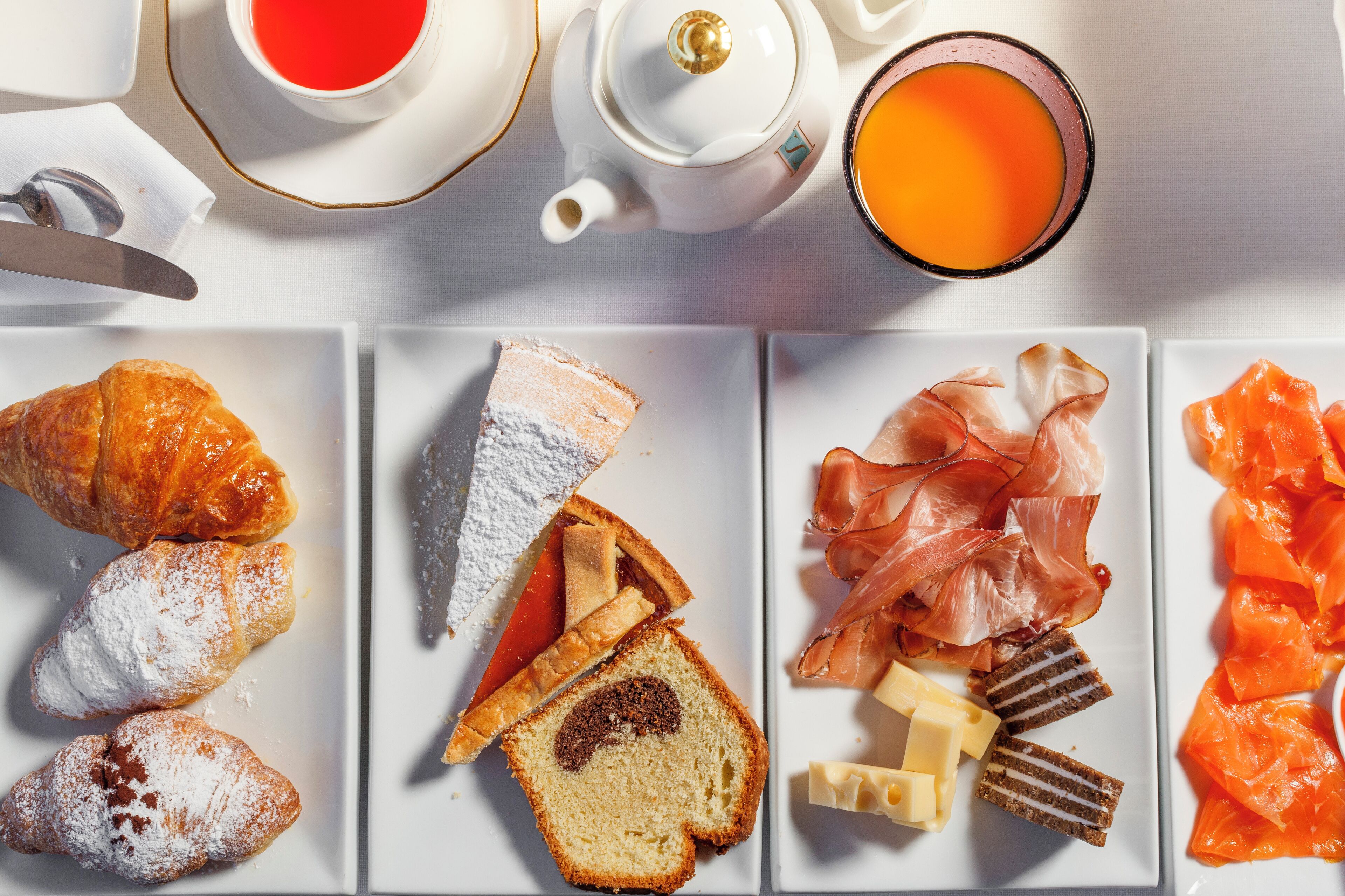 Ein Frühstücksbuffet mit Croissants, Puderzuckergebäck, Brot mit Marmelade, Räucherlachs, Käse und einem Teekessel mit einer Tasse Tee.