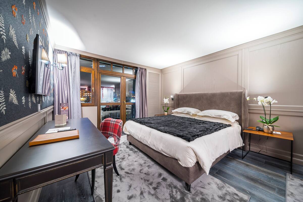 Ein Schlafzimmer mit Bett, Schreibtisch, Stuhl, Fernseher und einem Fenster mit Vorhängen.