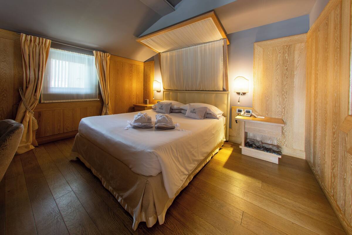 Ein ordentlich bezogenes Bett mit Kissen und Handtüchern, Holzboden, ein Nachttisch mit Lampe und ein Fenster mit Vorhängen.