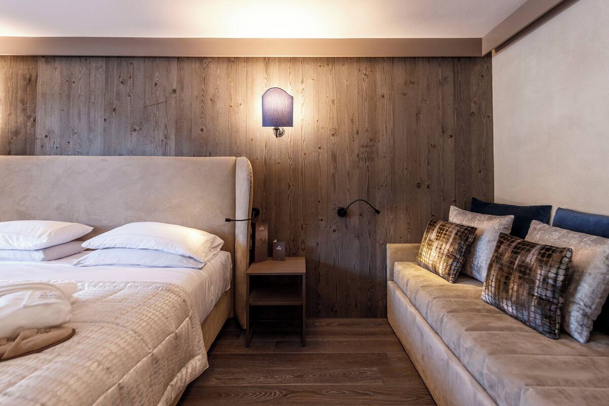 Ein Schlafzimmer mit Holzwand, einem Nachttisch, einem Bett mit weißer Bettwäsche und einer Couch mit gemusterten Kissen.