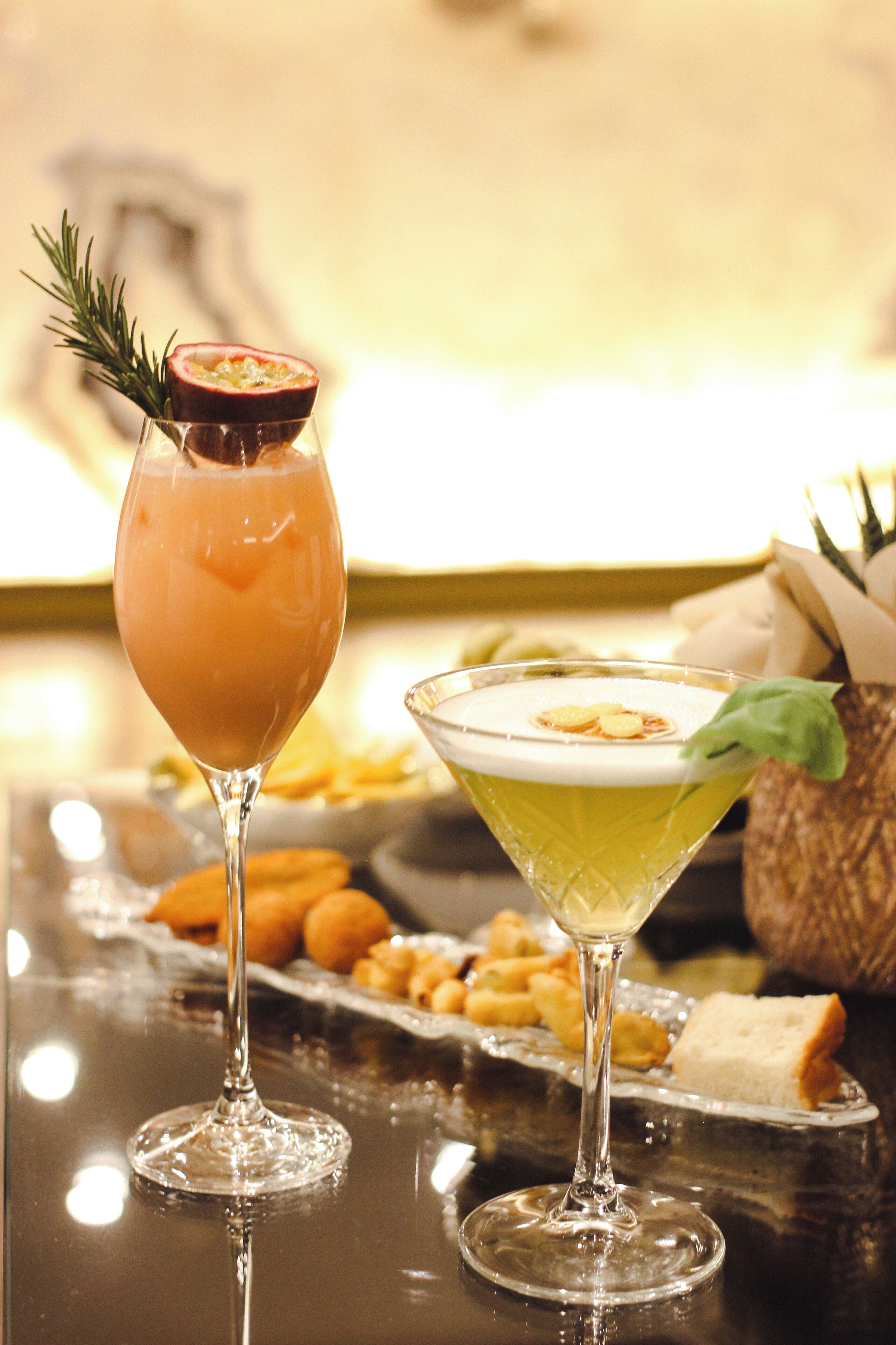 Zwei Cocktails auf einer spiegelnden Bar, begleitet von einer Vorspeisenplatte und einem Korb im Hintergrund.