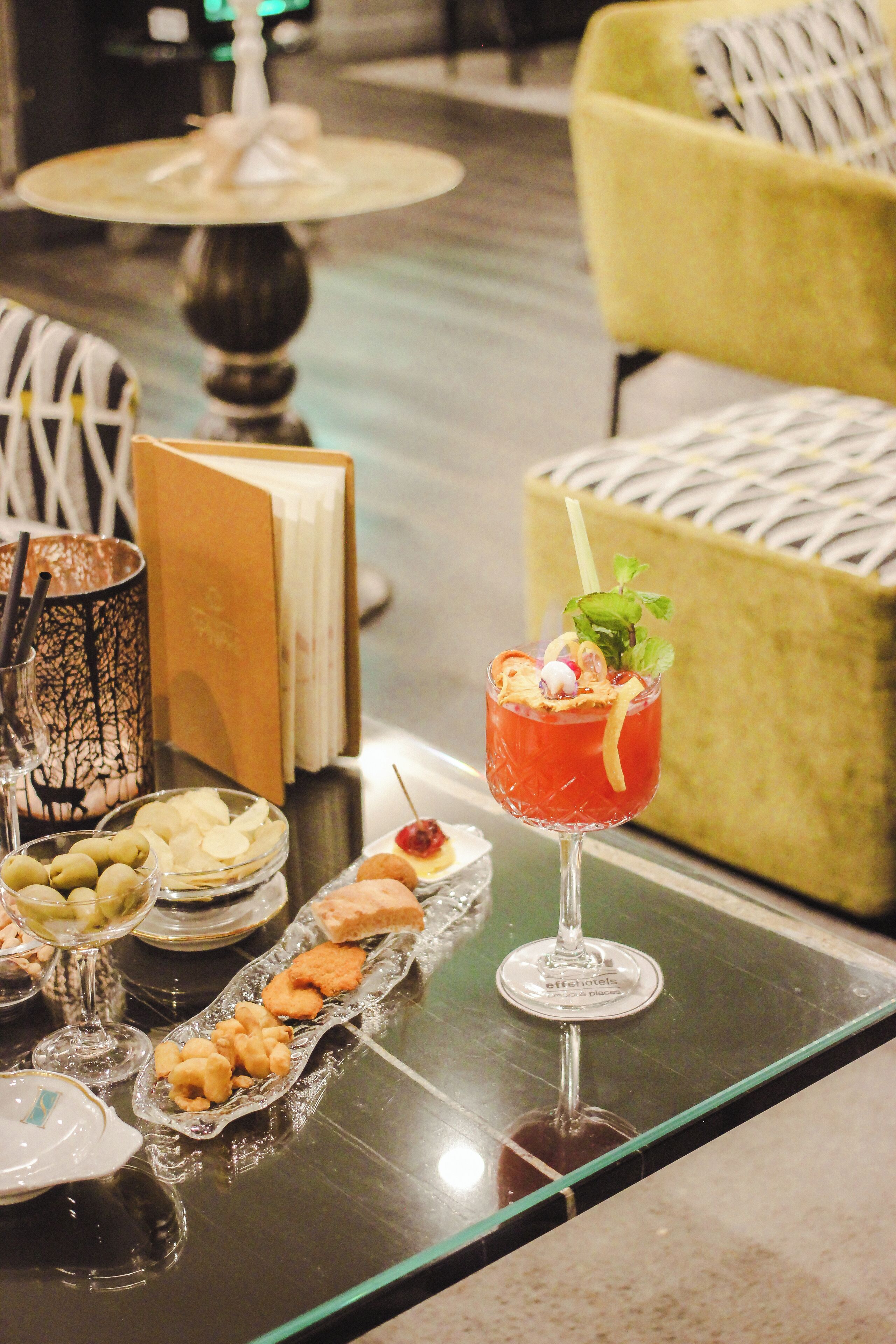 A table with a variety of food items, a glass of red cocktail, and a book.