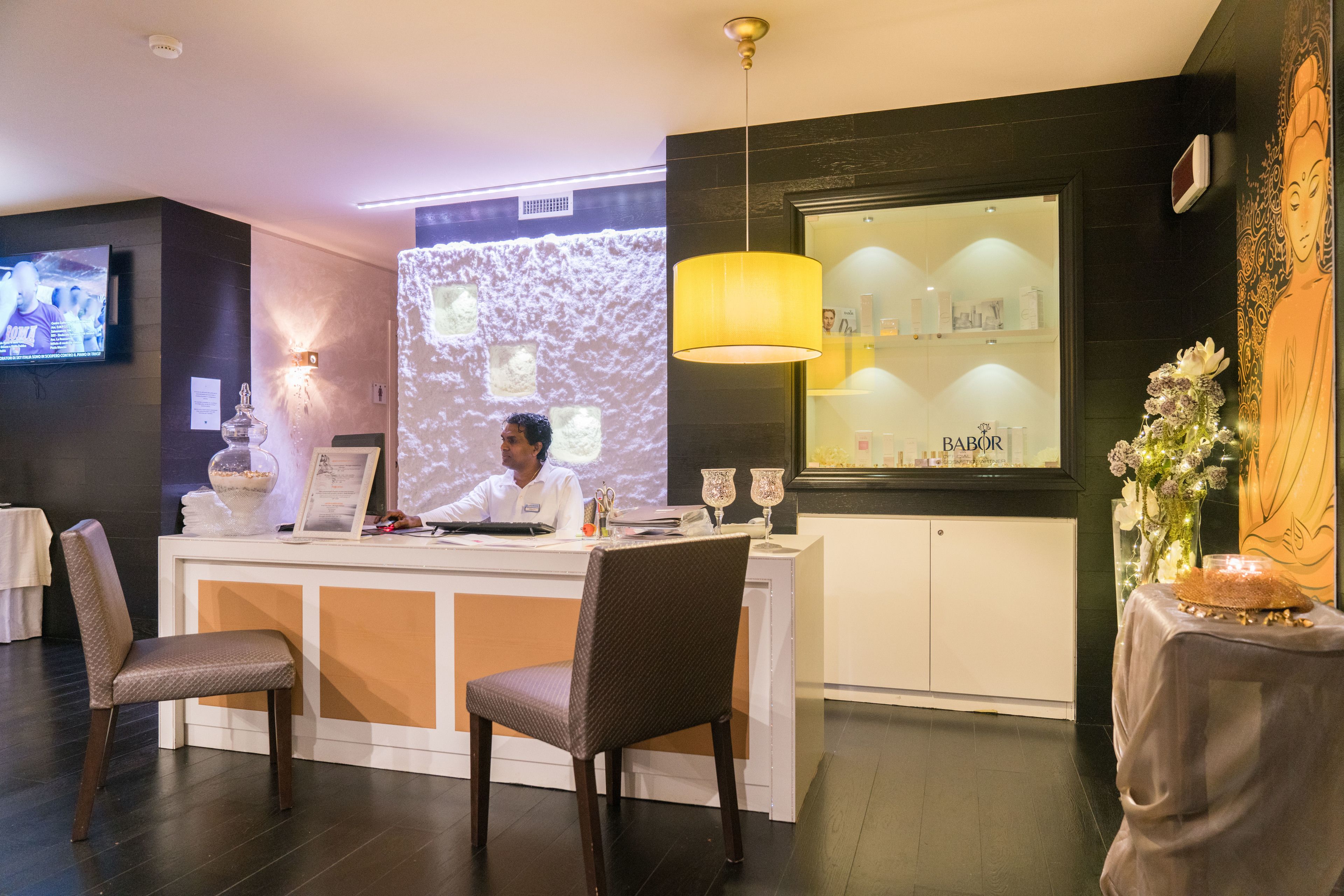 Zona reception di un hotel, con scrivania, sedute, schermo televisivo e parete con proiezione e mensole che espongono oggetti.