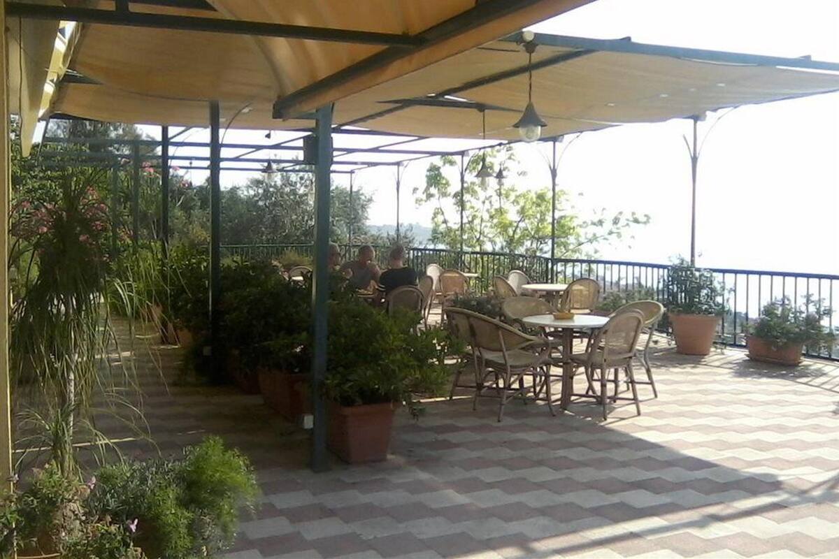 An outdoor patio with tables and chairs, surrounded by potted plants and a view of the sea.