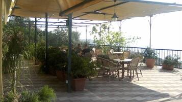 An outdoor patio with tables and chairs, surrounded by potted plants and a view of the sea.
