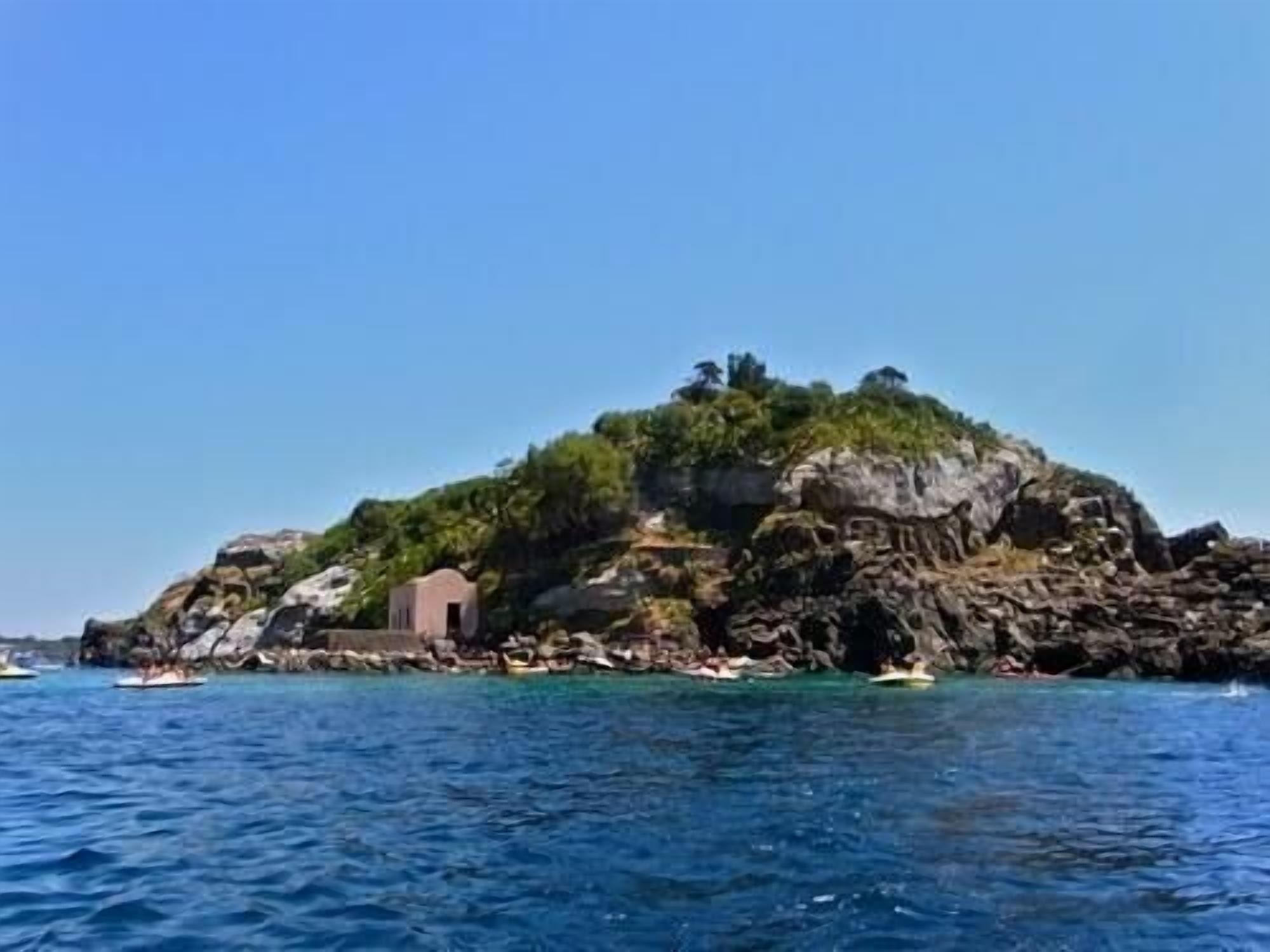 Un'isola rocciosa con un piccolo edificio, circondata da acque cristalline e imbarcazioni.