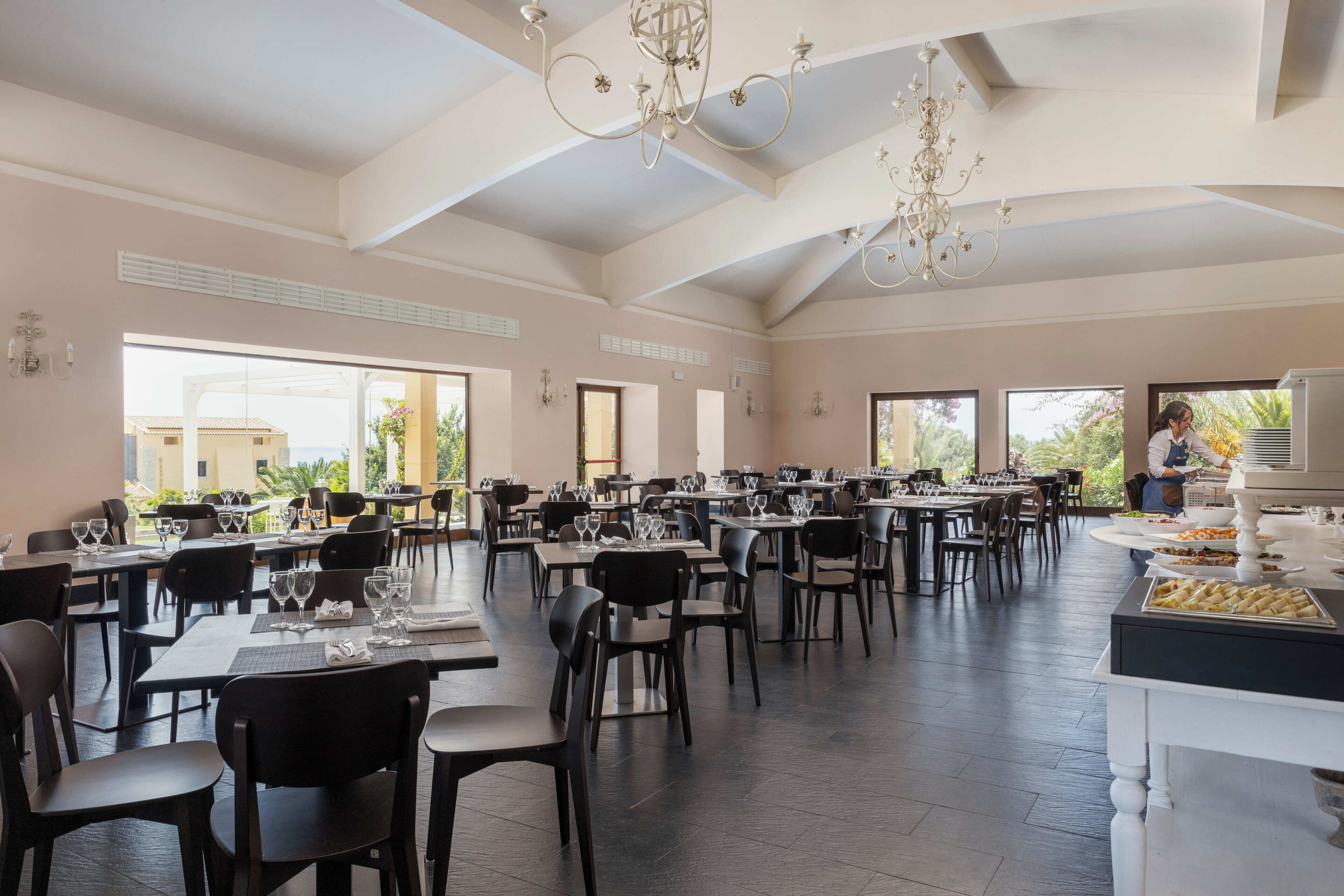 A spacious dining area with multiple tables and chairs, large windows, and a chandelier.