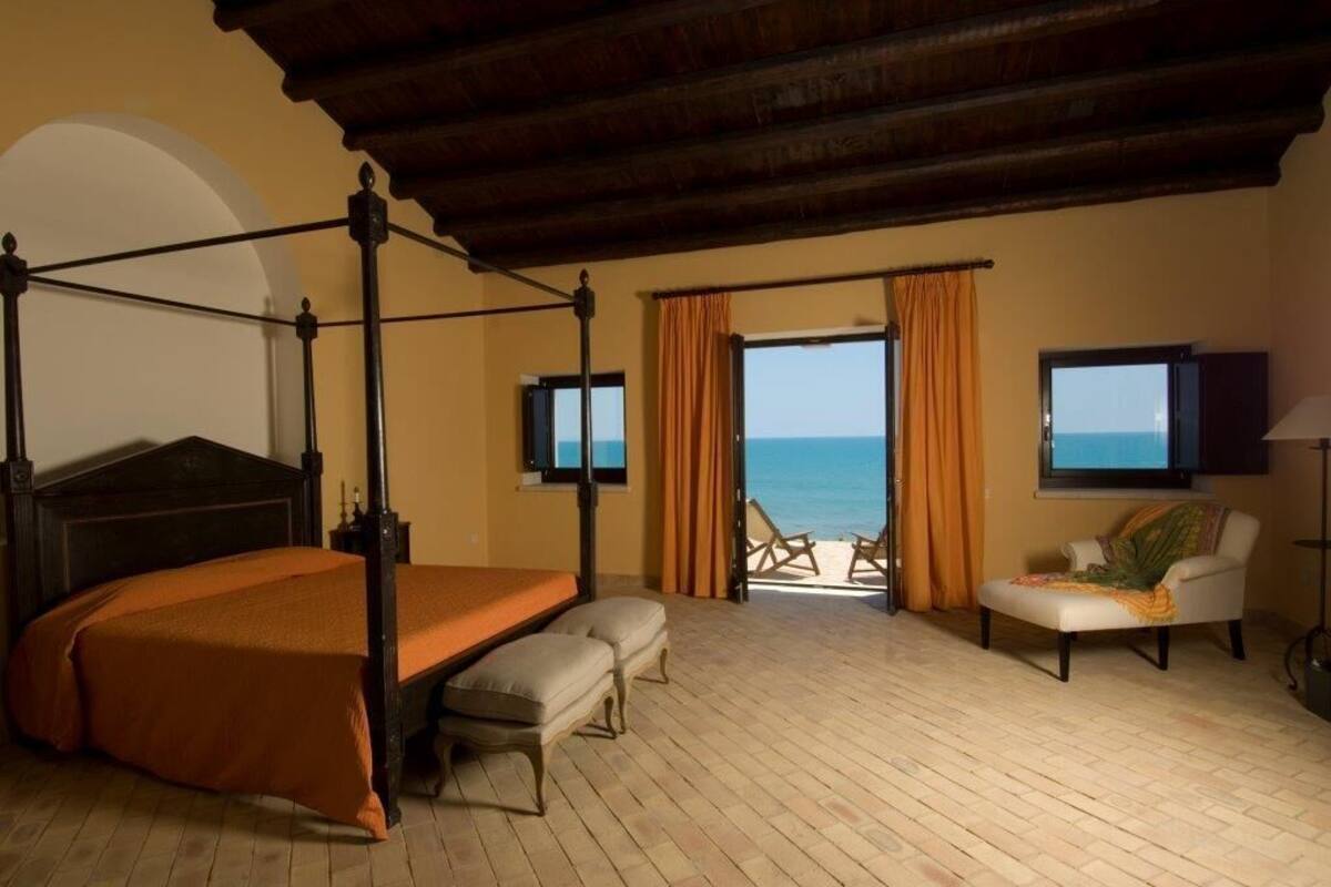 A bedroom with a four-poster bed, a balcony with a view of the sea, and a chair.