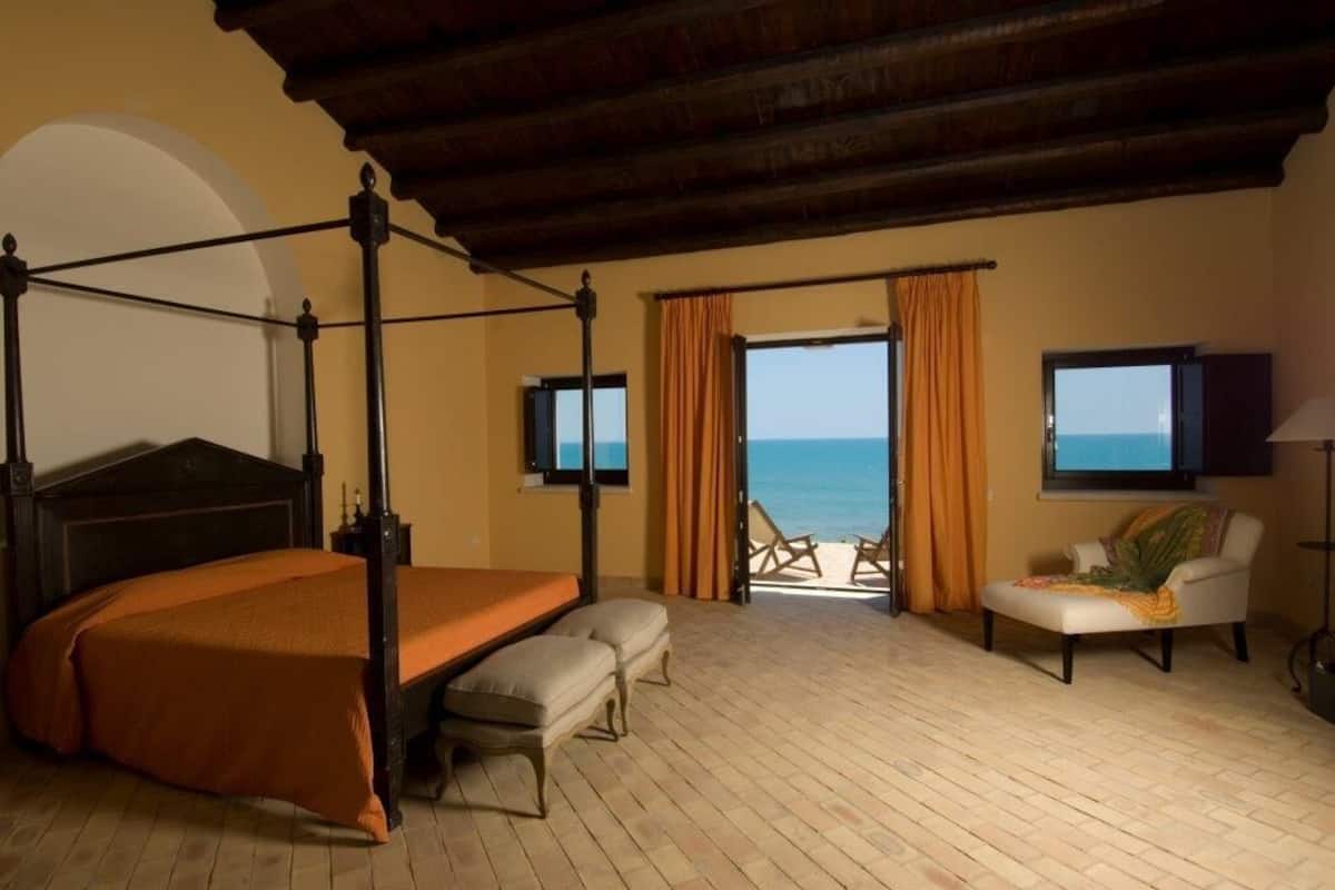 A bedroom with a four-poster bed, a balcony with a view of the sea, and a chair.