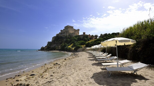 A beach with white sun loungers and umbrellas, a castle on a hill, and clear blue skies. - Falconara Greenblu Resort (Butera)