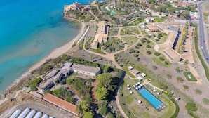 An aerial view of a coastal resort with a beach, villas, a swimming pool, and a large greenhouse area. - Falconara Greenblu Resort (Butera)