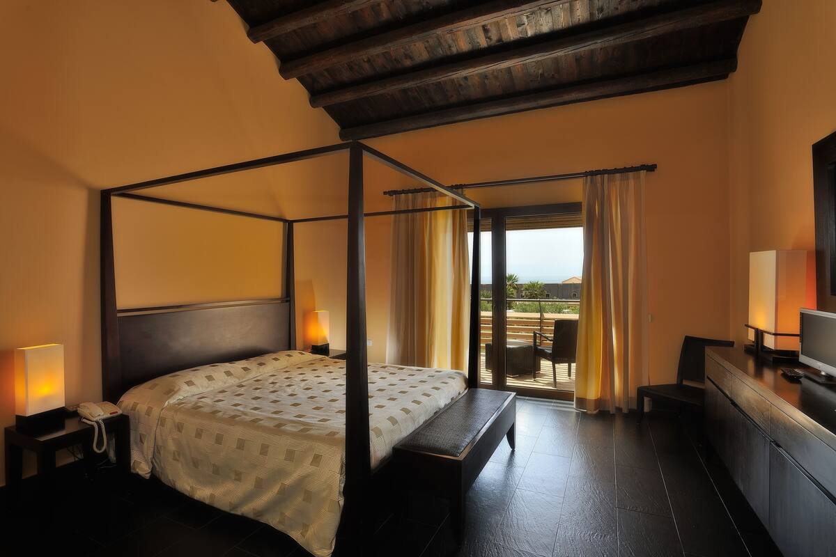 A four-poster bed with a canopy, bedside lamps, a desk with a television, and a view of the outdoors through a glass door.