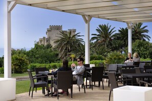 An outdoor patio with tables and chairs, a castle in the background, and palm trees. - Falconara Greenblu Resort (Butera)
