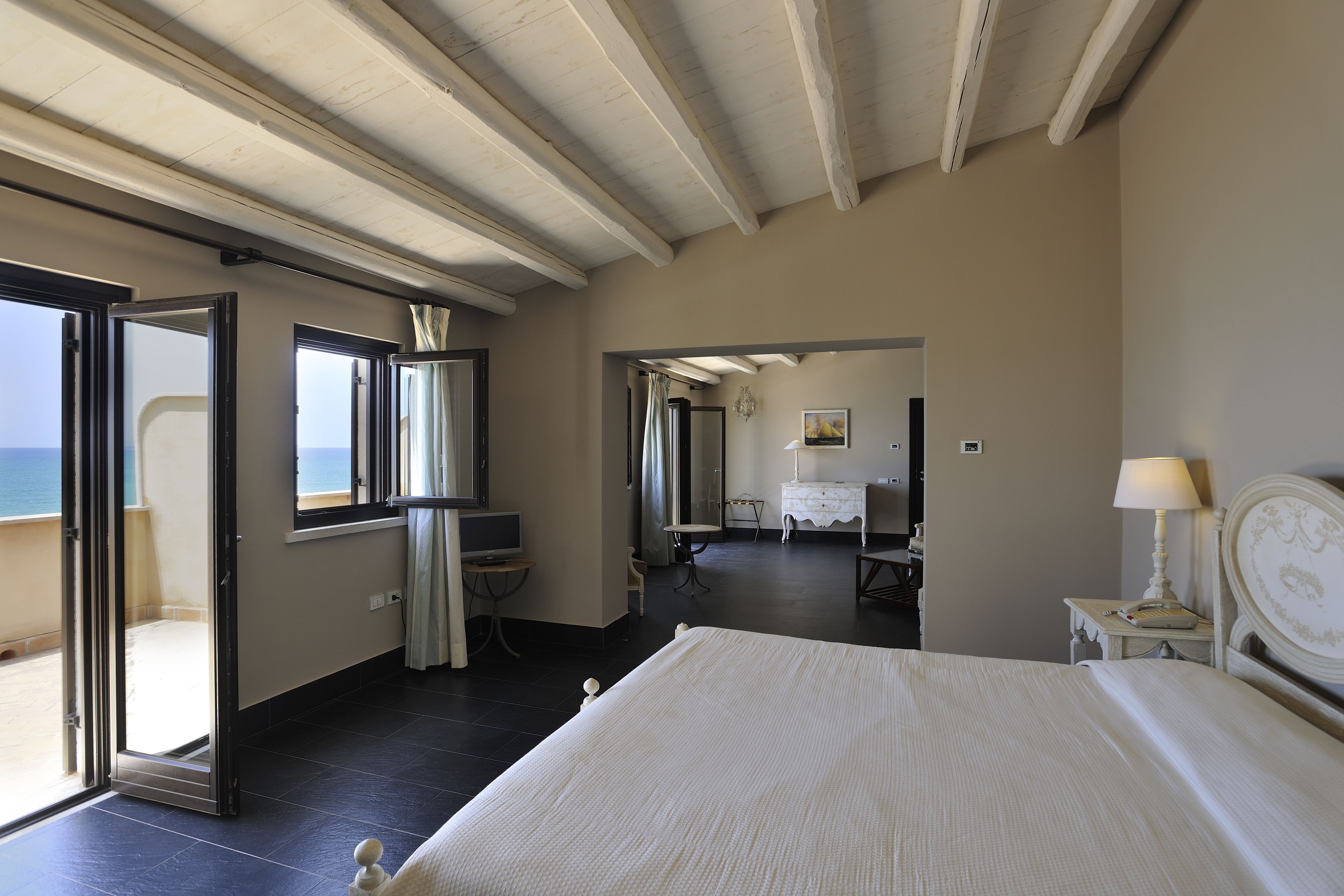 Quarto de hotel com cama, escrivaninha, cadeira, cômoda e vista para o mar por uma porta aberta.